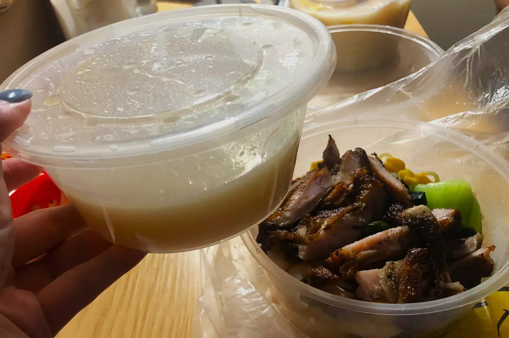 A hand holds a plastic container of broth above another container with grilled meat and vegetables on noodles, set on a wooden table, suggesting a warm meal.