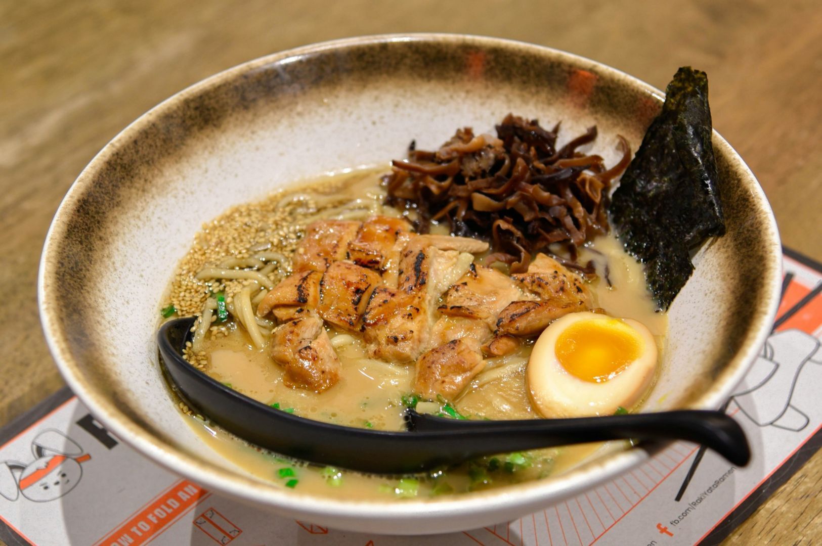 A steaming bowl of ramen with slices of char-grilled chicken, half a soft-boiled egg, a sheet of nori, brown mushrooms, sesame seeds, and chopped green onions resting in a rich, creamy broth.
