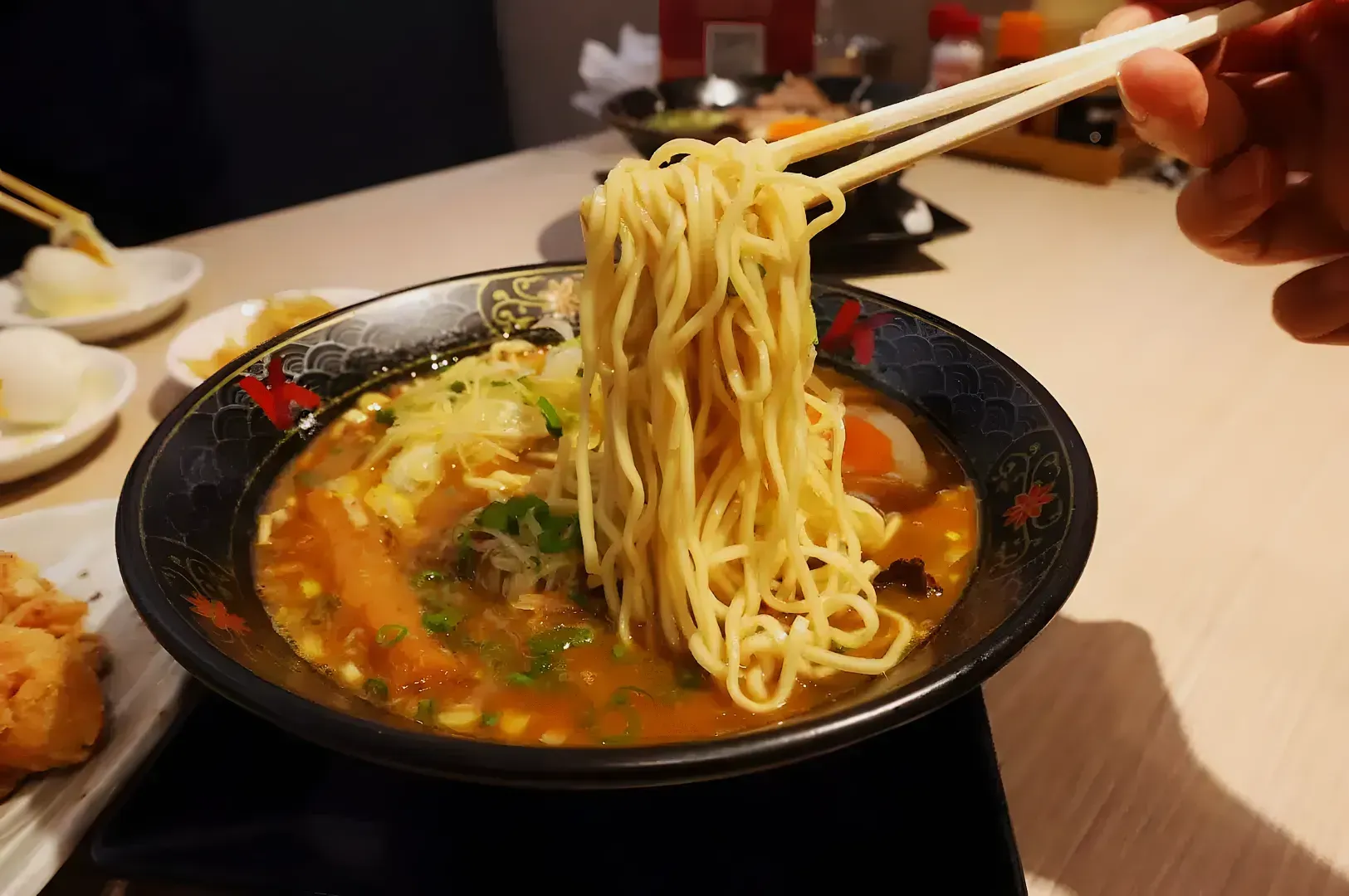 A bowl of ramen with chopsticks lifting noodles from rich broth. Toppings include sliced green onions and eggs. Warm, inviting dining scene.