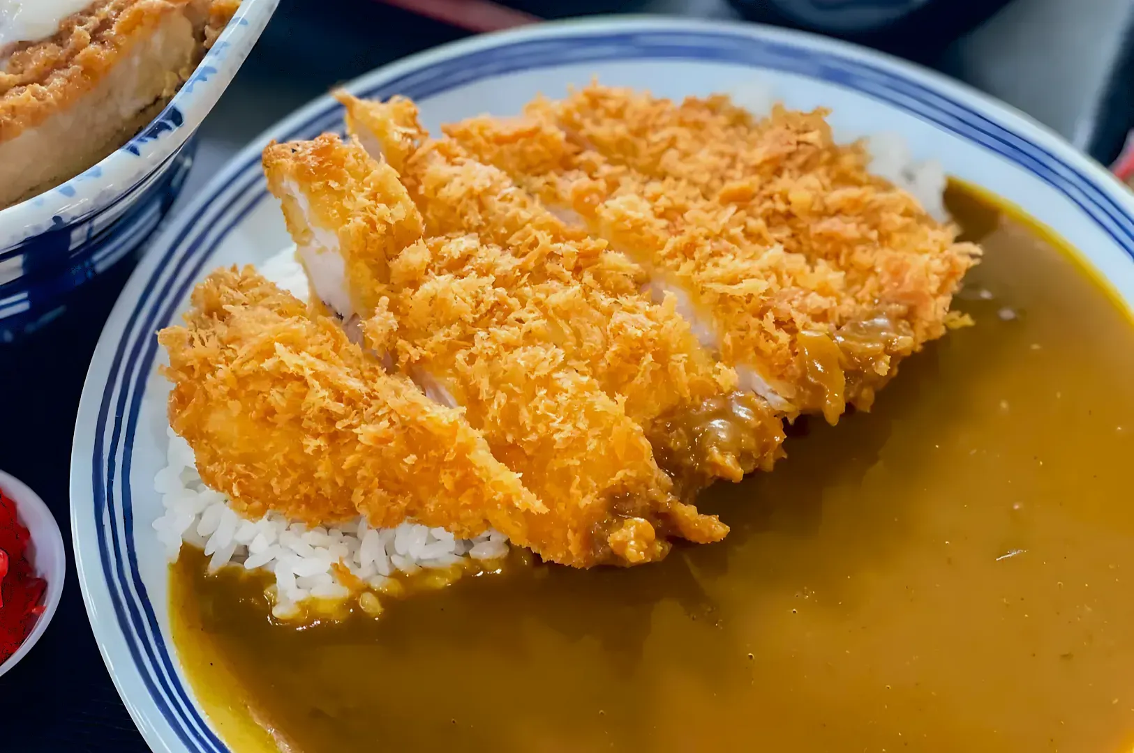 Crispy golden chicken katsu over rice, partially covered in rich brown curry sauce, on a white plate. A warm, hearty meal, invitingly presented.