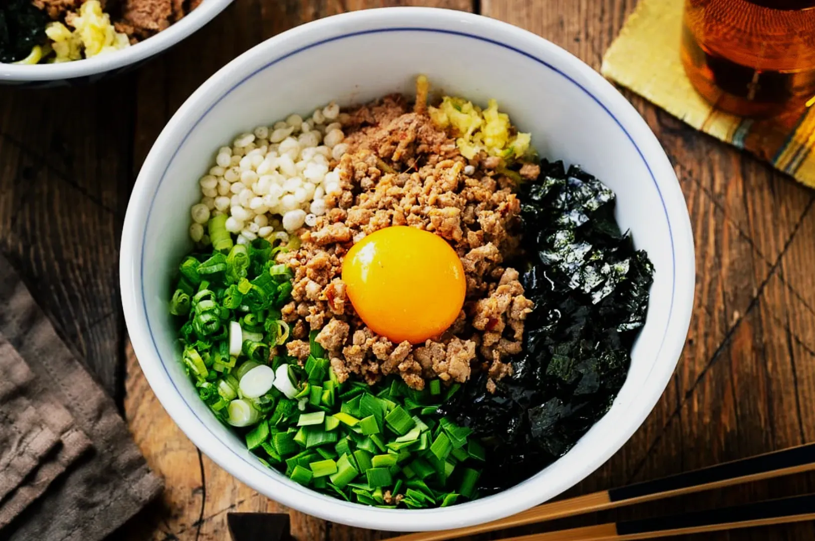 A bowl of Mazesoba with a raw egg yolk at the center, surrounded by minced meat, chopped green onions, nori, tempura bits, and ginger, on a wooden table.