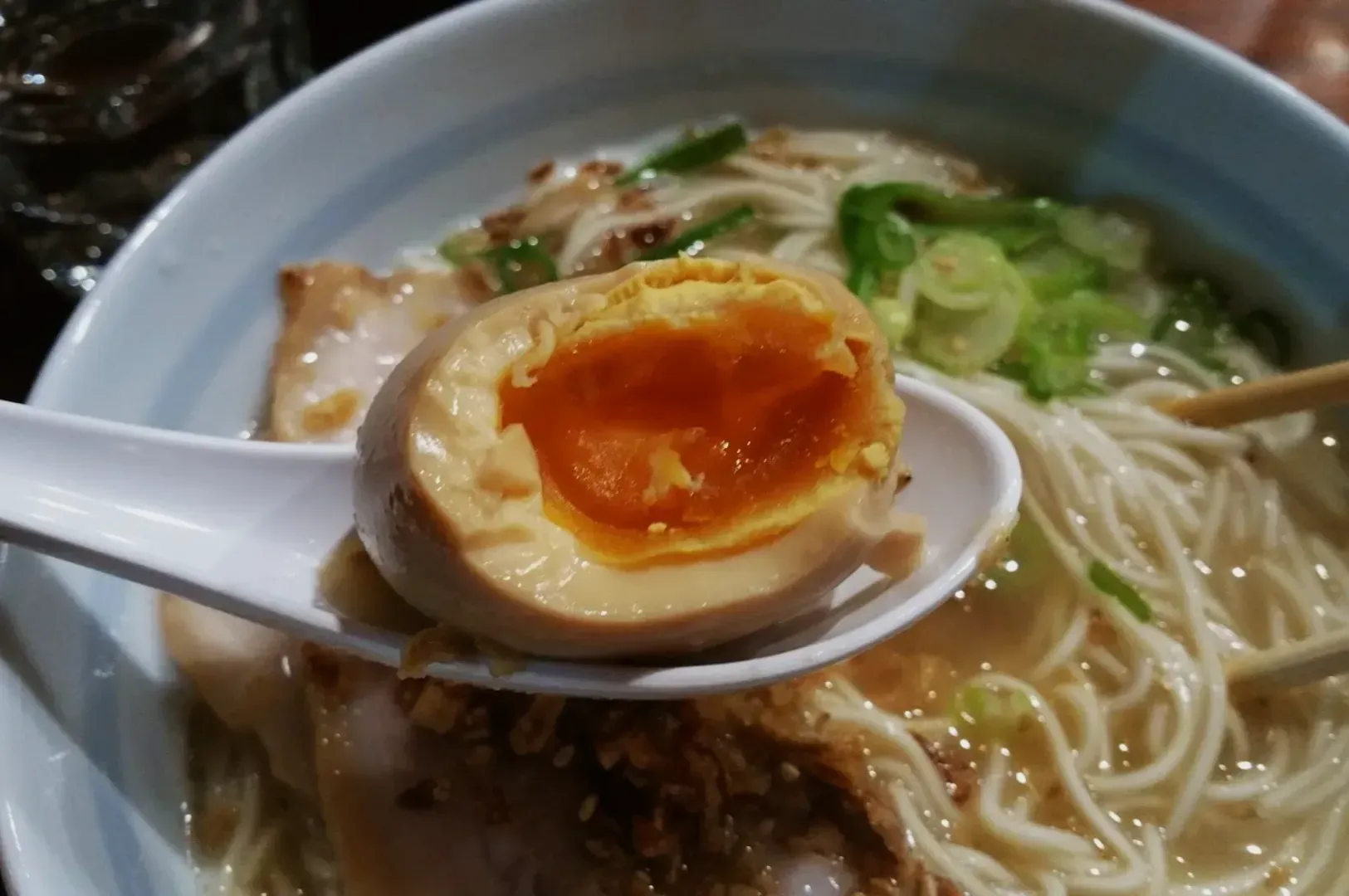 A bowl of ramen with noodles and green onions, topped with a sliced soft-boiled egg on a spoon, showcasing its golden yolk.