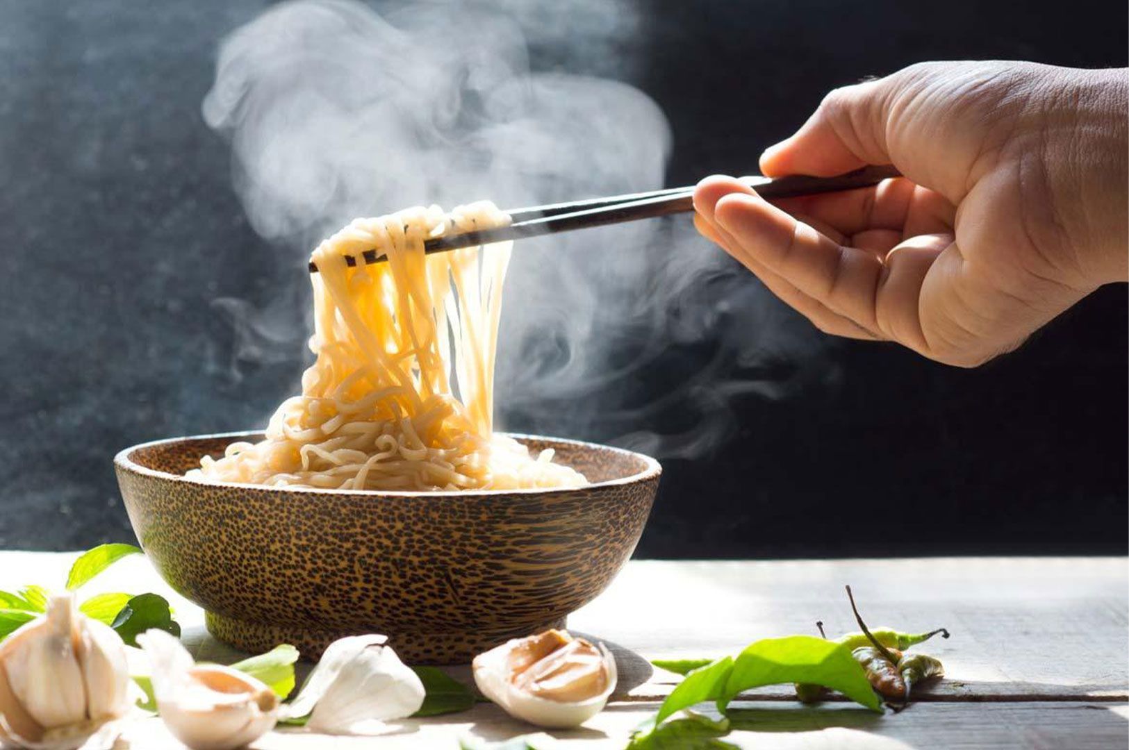 Close-up of a hand lifting steaming instant noodles from a wooden bowl with dark chopsticks, surrounded by fresh garlic cloves and green herbs.
