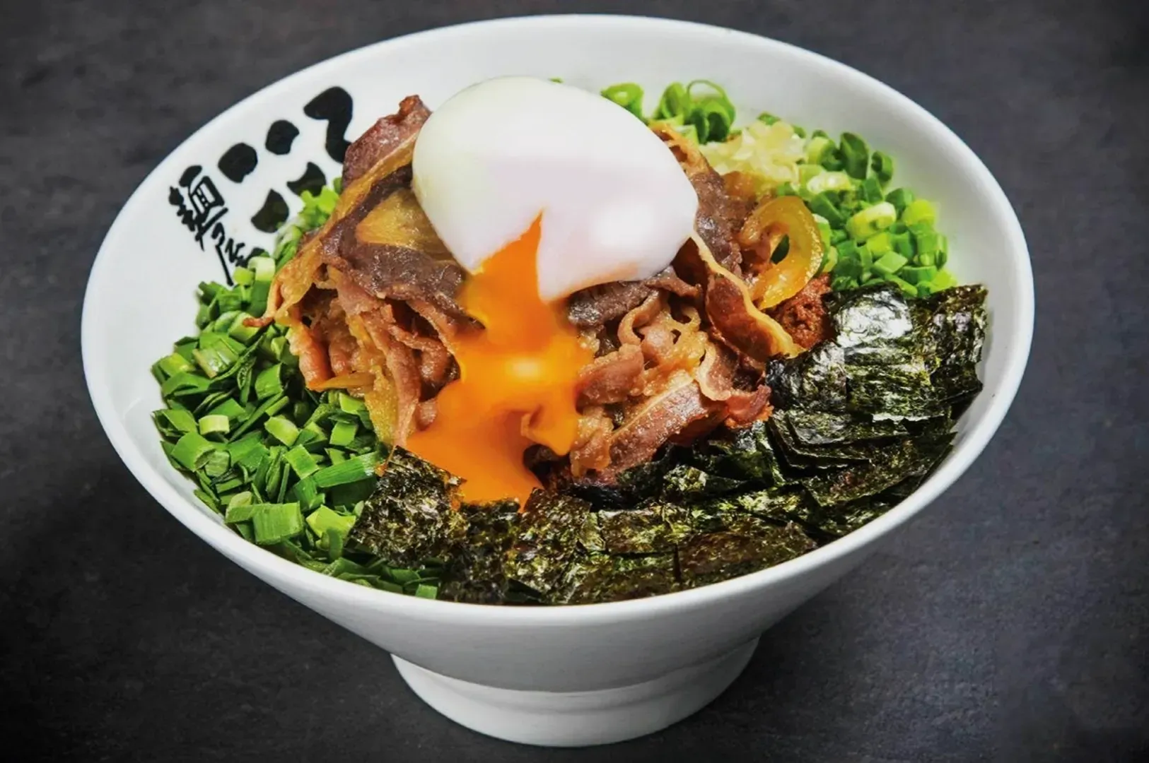 A bowl of ramen topped with a soft-boiled egg with runny yolk, green onions, sliced beef, and seaweed on a dark surface. The dish appears savory and appetizing.
