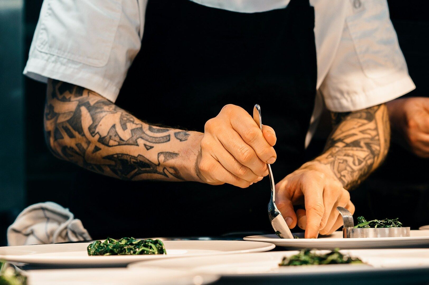 Chef with tattooed arms skillfully plates greens on dishes. The focus is on the precise, artistic food presentation, conveying professionalism and creativity.