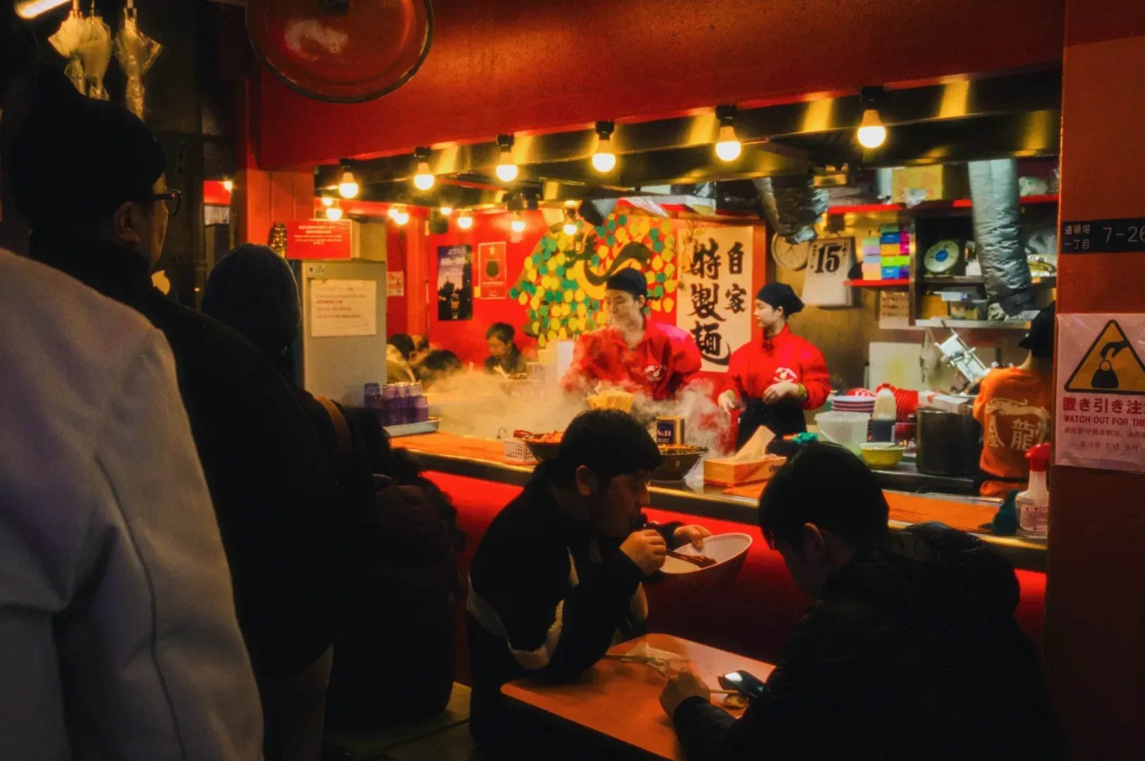 A bustling ramen shop with chefs in red uniforms cooking over steaming pots. Customers sit at a counter, enjoying their meals. Warm, cozy ambiance.