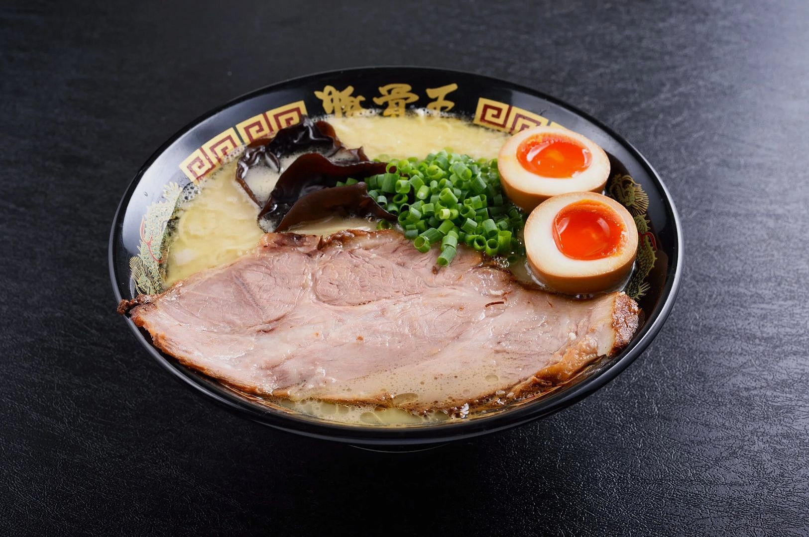 A front view of a rich ramen bowl with a large pork slice, two halved soft-boiled eggs, a mound of chopped green onions, and dark wood ear mushrooms in a creamy broth.