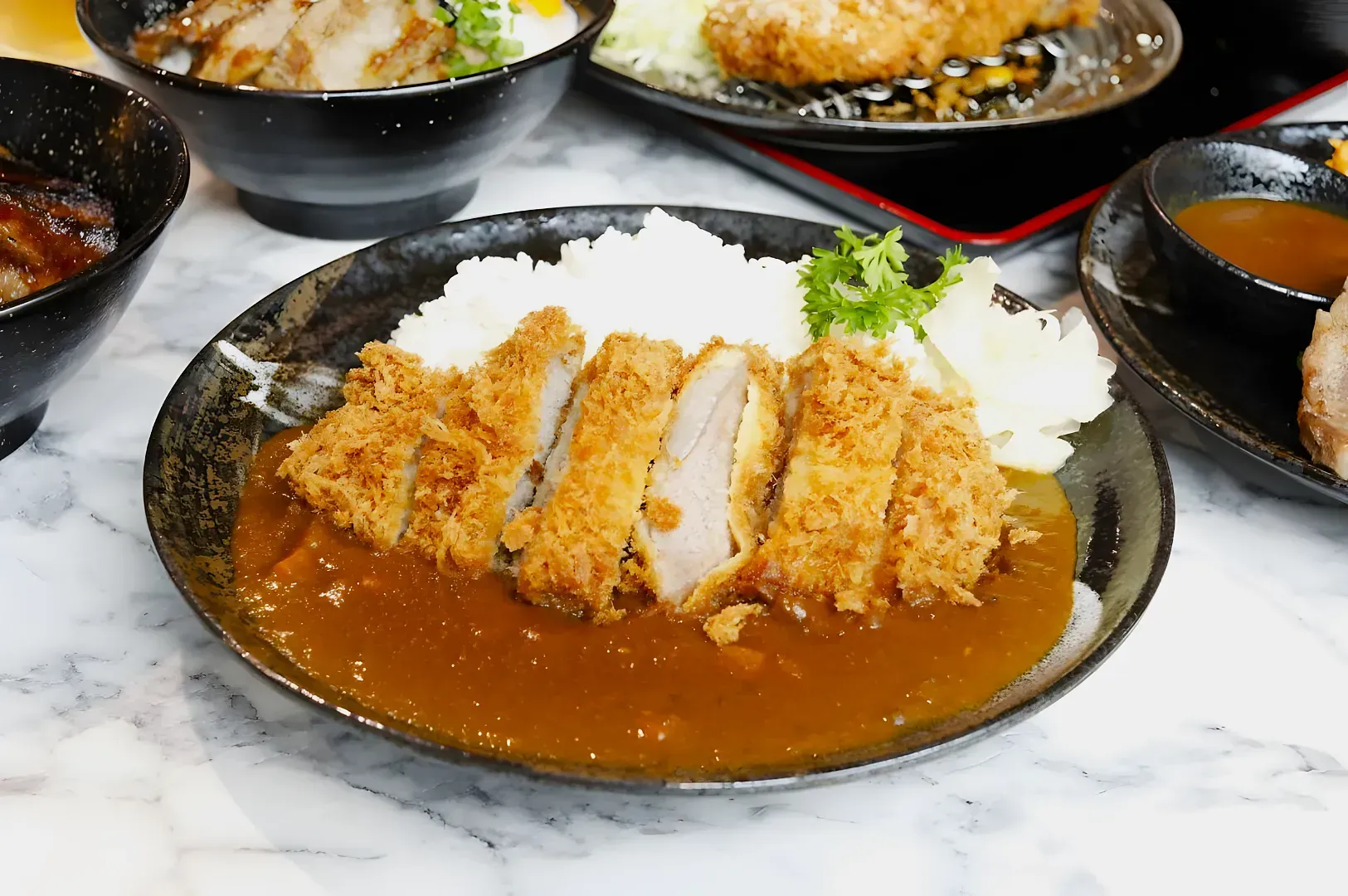 A plate of Japanese curry with sliced, crispy breaded pork cutlet on top, served with white rice and a garnish of parsley on a marble table.