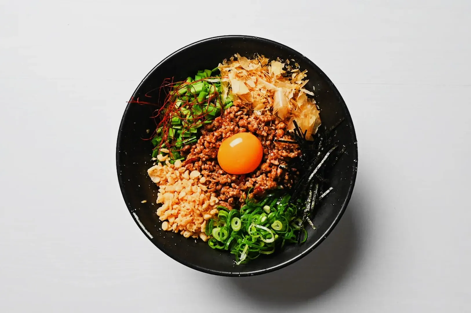 A black bowl filled with colorful ingredients: minced meat, a raw egg yolk in the center, green onions, seaweed strips, and bonito flakes.