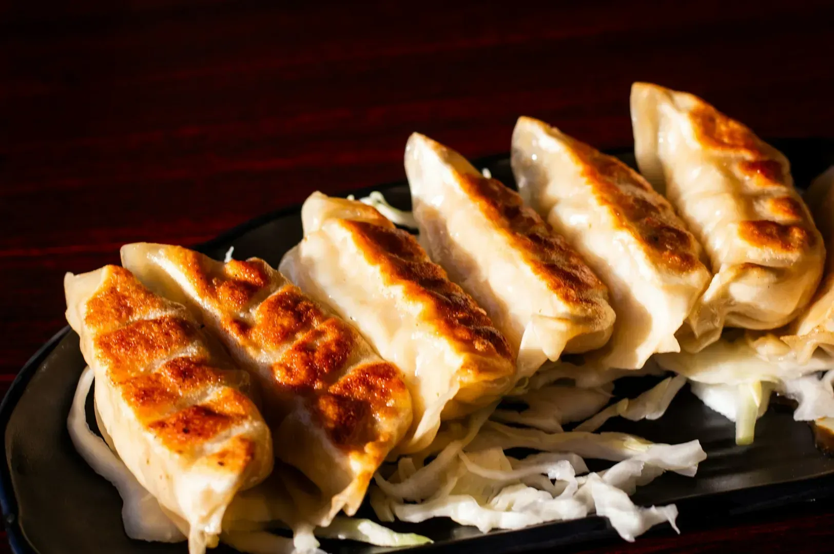 Golden-brown pan-fried dumplings are neatly arranged on a dark plate over shredded cabbage, creating a warm, appetizing display against a rich wood background.