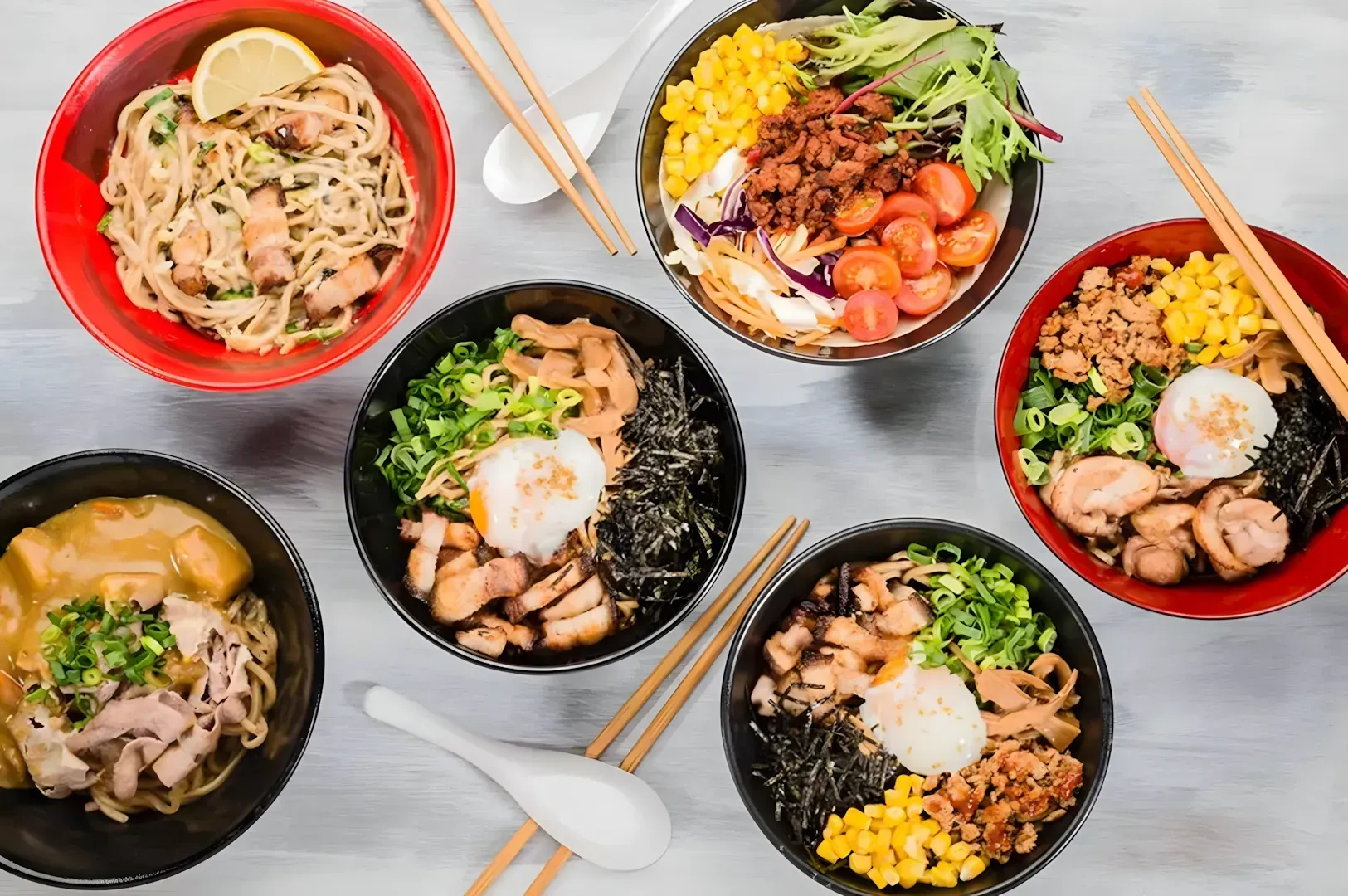 Aerial view of six colorful bowls of ramen, each garnished with ingredients like corn, tomatoes, and green onions, alongside chopsticks and spoons.