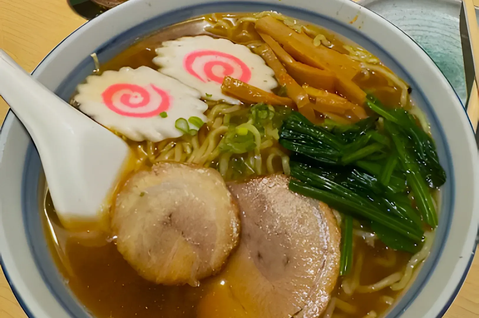 A bowl of ramen with slices of chashu pork, narutomaki with pink spirals, bamboo shoots, greens, and noodles in broth. A spoon rests on the bowl's edge.