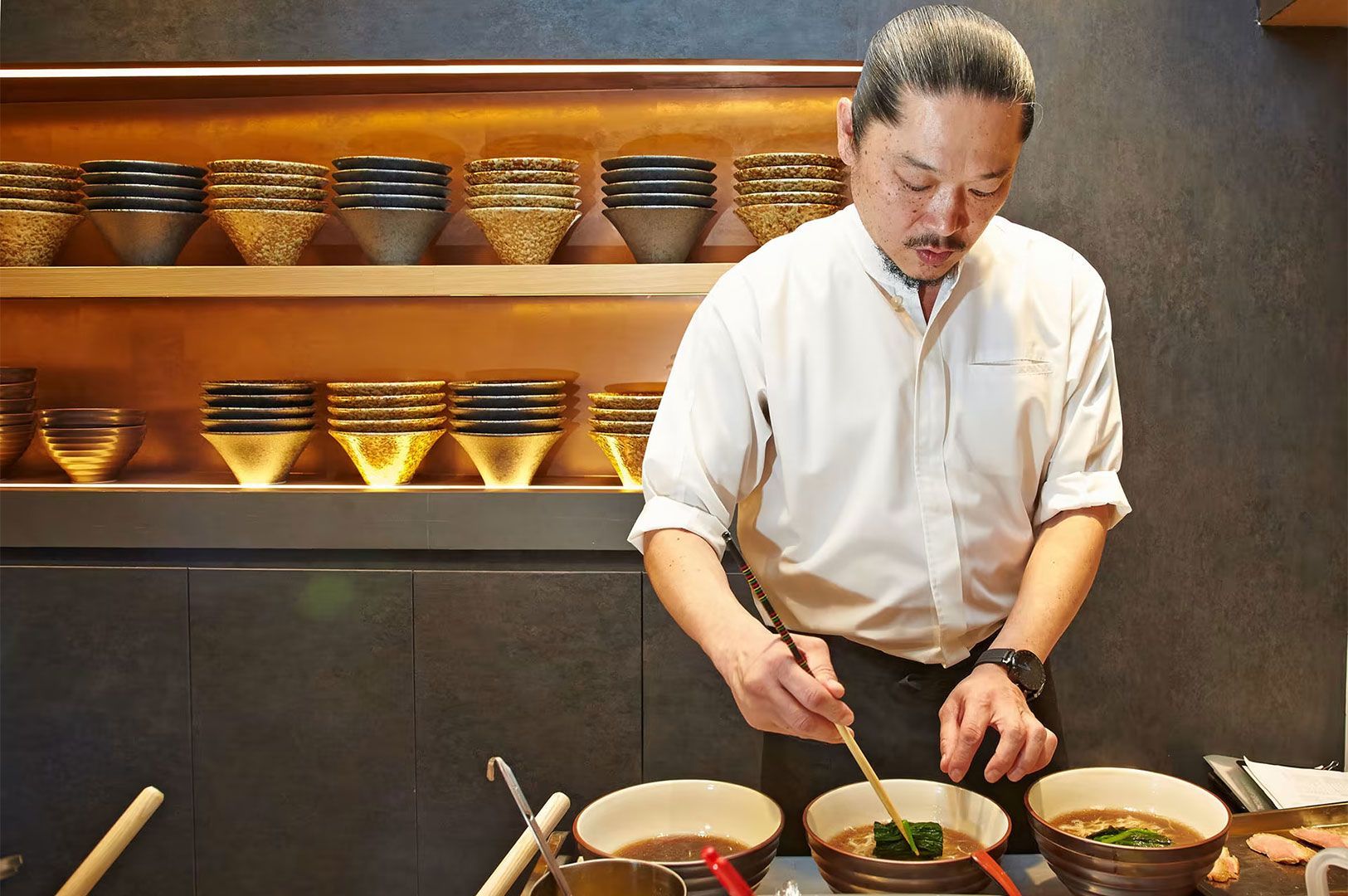 A Japanese chef in a white shirt and apron carefully garnishing bowls of ramen on a counter, with stacked ceramic bowls displayed on shelves behind him.