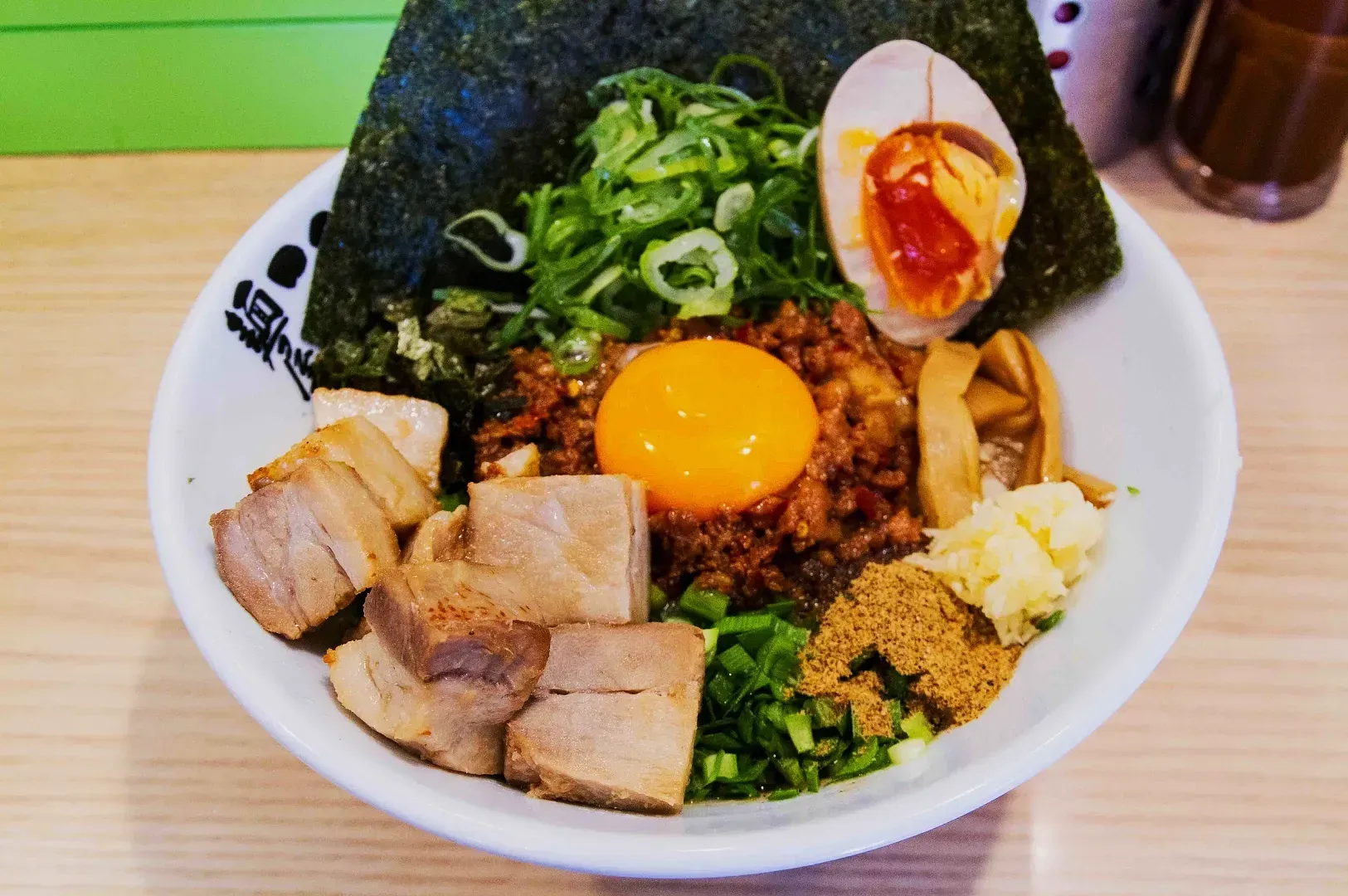 A bowl of ramen topped with a raw egg yolk, char siu pork cubes, seaweed, green onions, a spicy egg, bamboo shoots, and minced garlic.