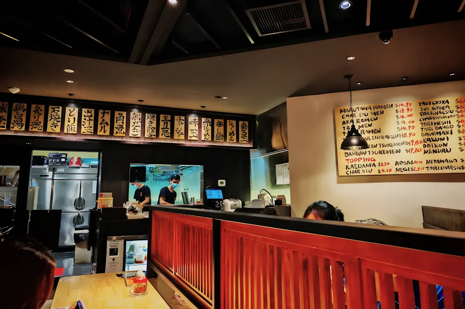 Cozy Japanese restaurant interior with wooden details and wall menu boards featuring Japanese and English text. Chefs in the kitchen, creating a busy, welcoming atmosphere.