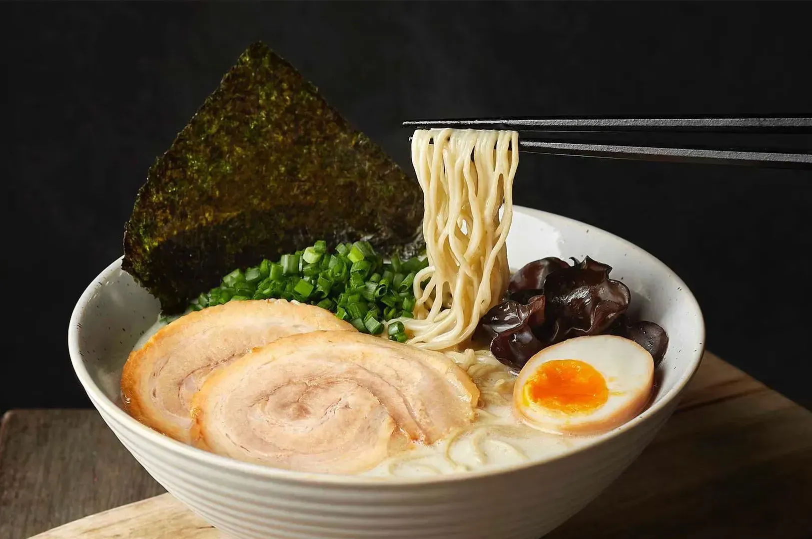 A steaming bowl of ramen with chopsticks holding noodles. Toppings include sliced pork, boiled egg, seaweed, mushrooms, and green onions. Cozy and appetizing.
