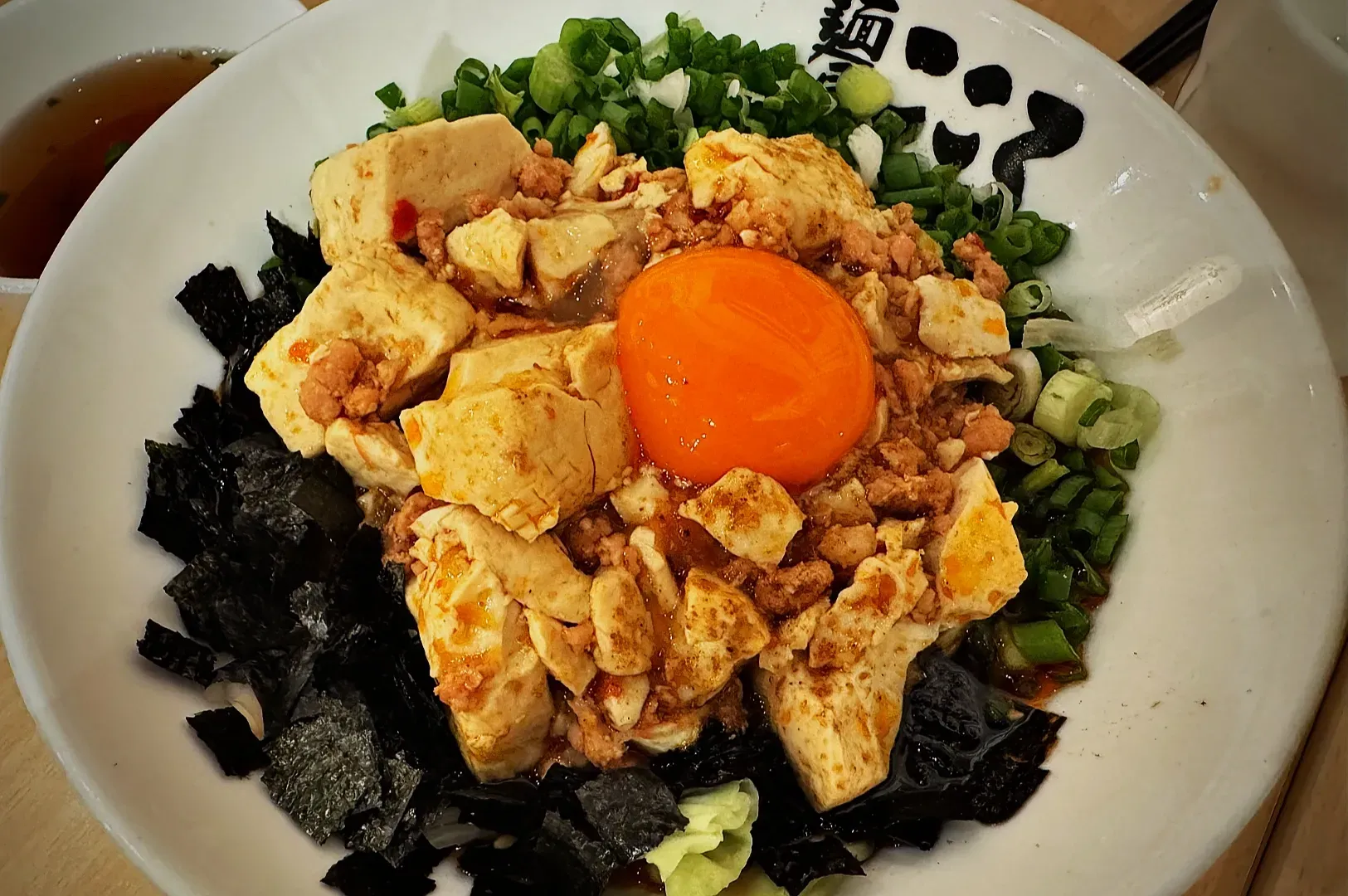 A vibrant bowl of noodles topped with creamy tofu, minced meat, a bright orange egg yolk, fresh chopped green onions, and dark seaweed strips. The dish looks savory and appetizing.