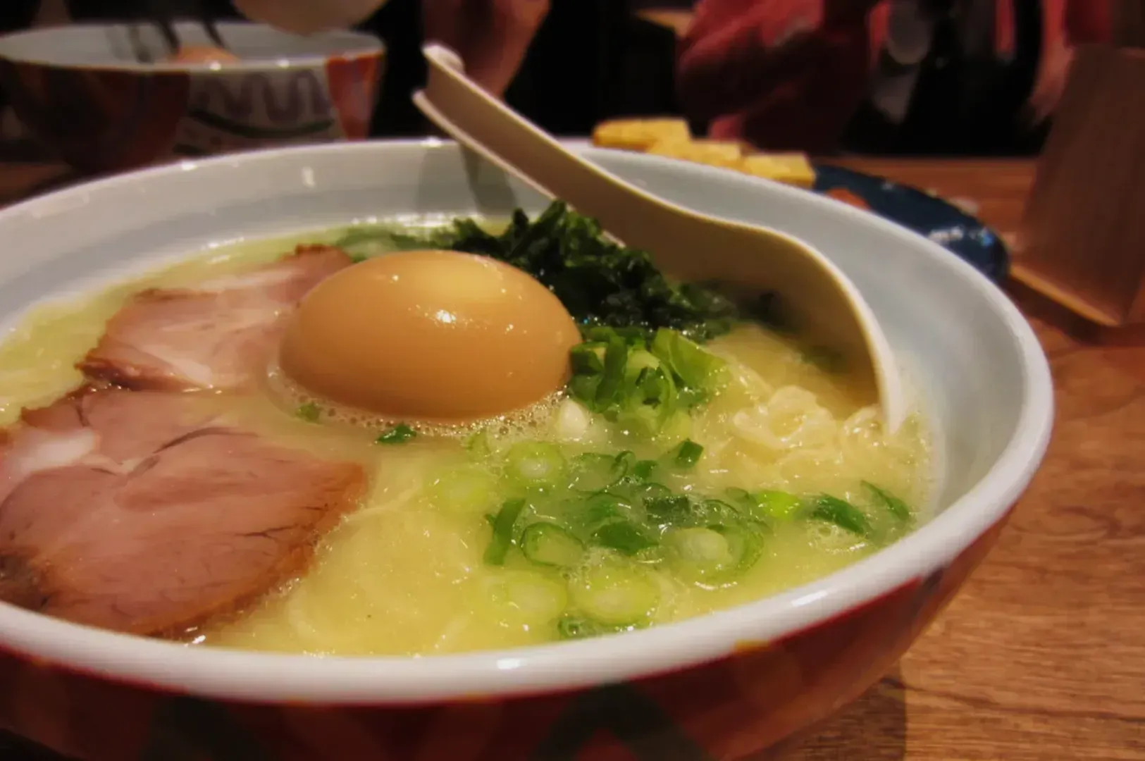A steaming bowl of ramen with slices of pork, a soft-boiled egg, green onions, and seaweed in a flavorful broth, set on a wooden table.