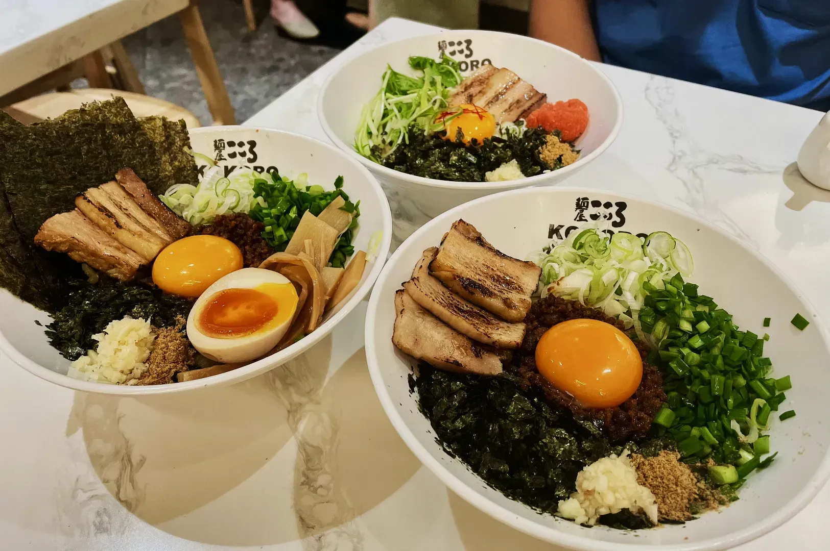 Three bowls of ramen on a marble table, each featuring a raw egg yolk, seaweed, green onions, grilled pork, and a seasoned soft-boiled egg.