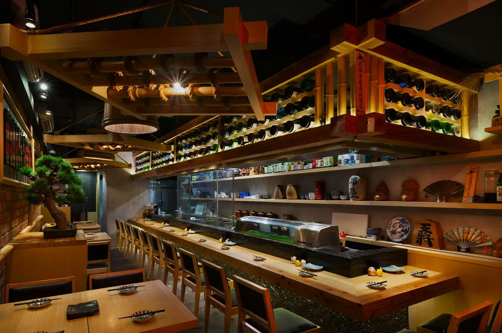 Cozy sushi bar with wooden decor, soft lighting, and a bonsai tree. Neatly arranged sushi counter with chairs, shelves of bottles, and Japanese pottery.