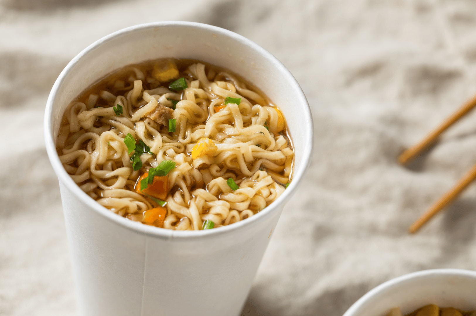 A disposable cup of instant ramen garnished with carrots and green onions, set against a soft beige background. Though simple and quick, this humble bowl invites fermentation-forward enhancements—like kimchi, pickled radish, or miso paste—to elevate its broth and balance its saltiness with tang and depth.
