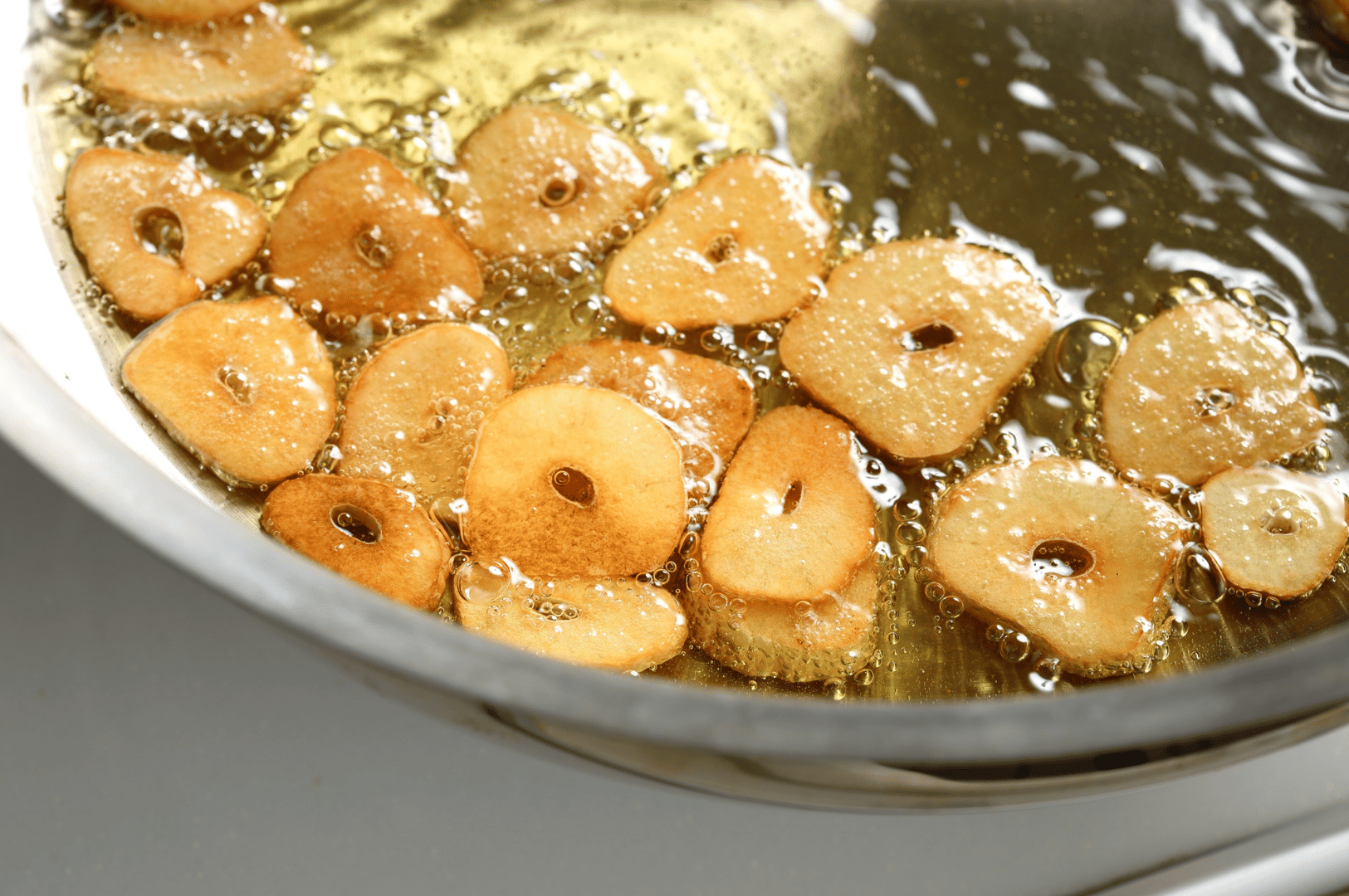 Close-up of thin garlic slices sizzling in hot oil, turning golden brown as bubbles rise around them—capturing the precise moment when raw pungency transforms into aromatic crispness. This foundational step in garlic infusion sets the flavor base for a deeply layered ramen broth, evoking both technique and sensory anticipation.