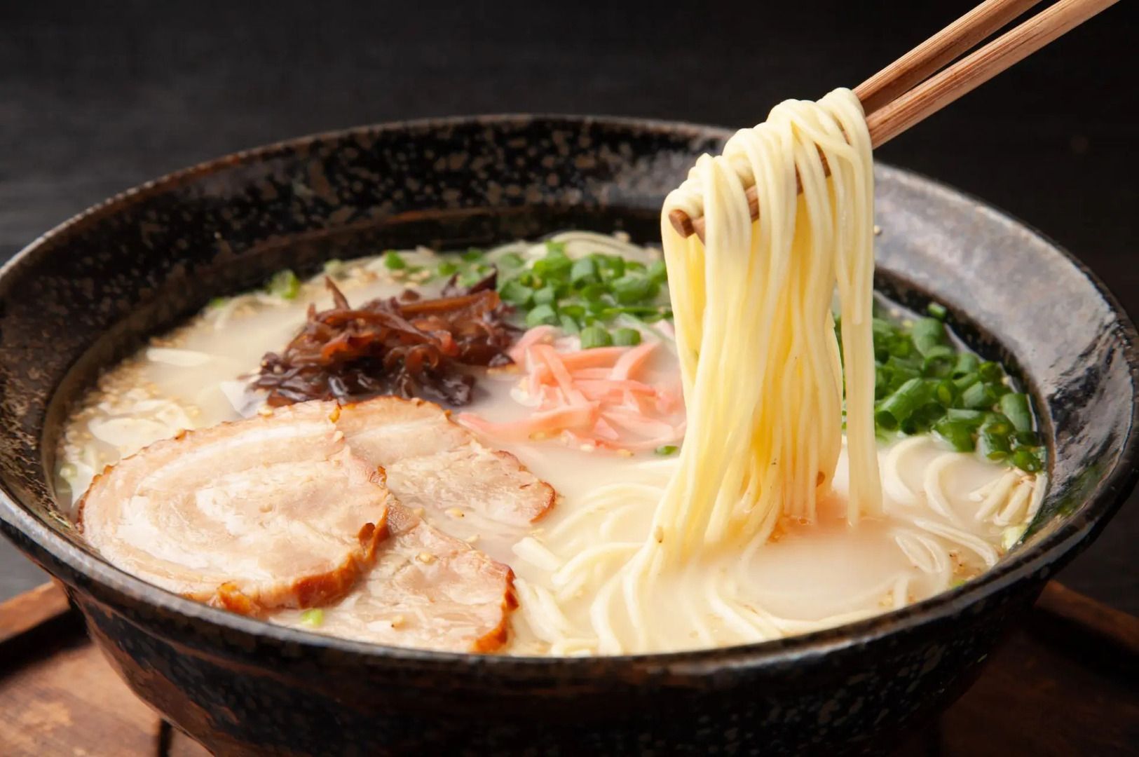 A serving of Hakata Tonkotsu Ramen featuring meat, with chopsticks picking noodles from the bowl.
