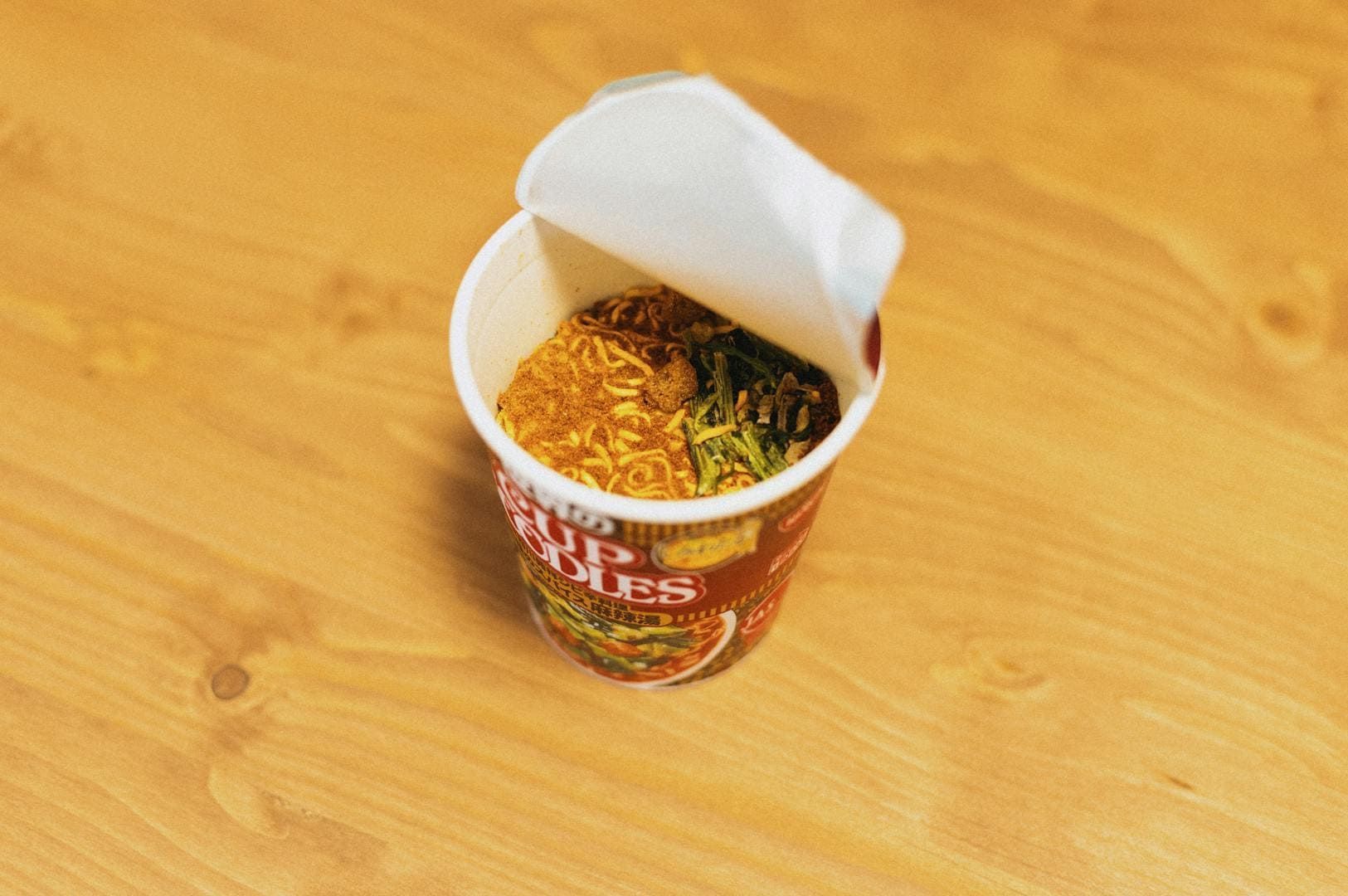 A sealed cup of instant noodles placed on top of a wooden table, awaiting hot water for cooking.