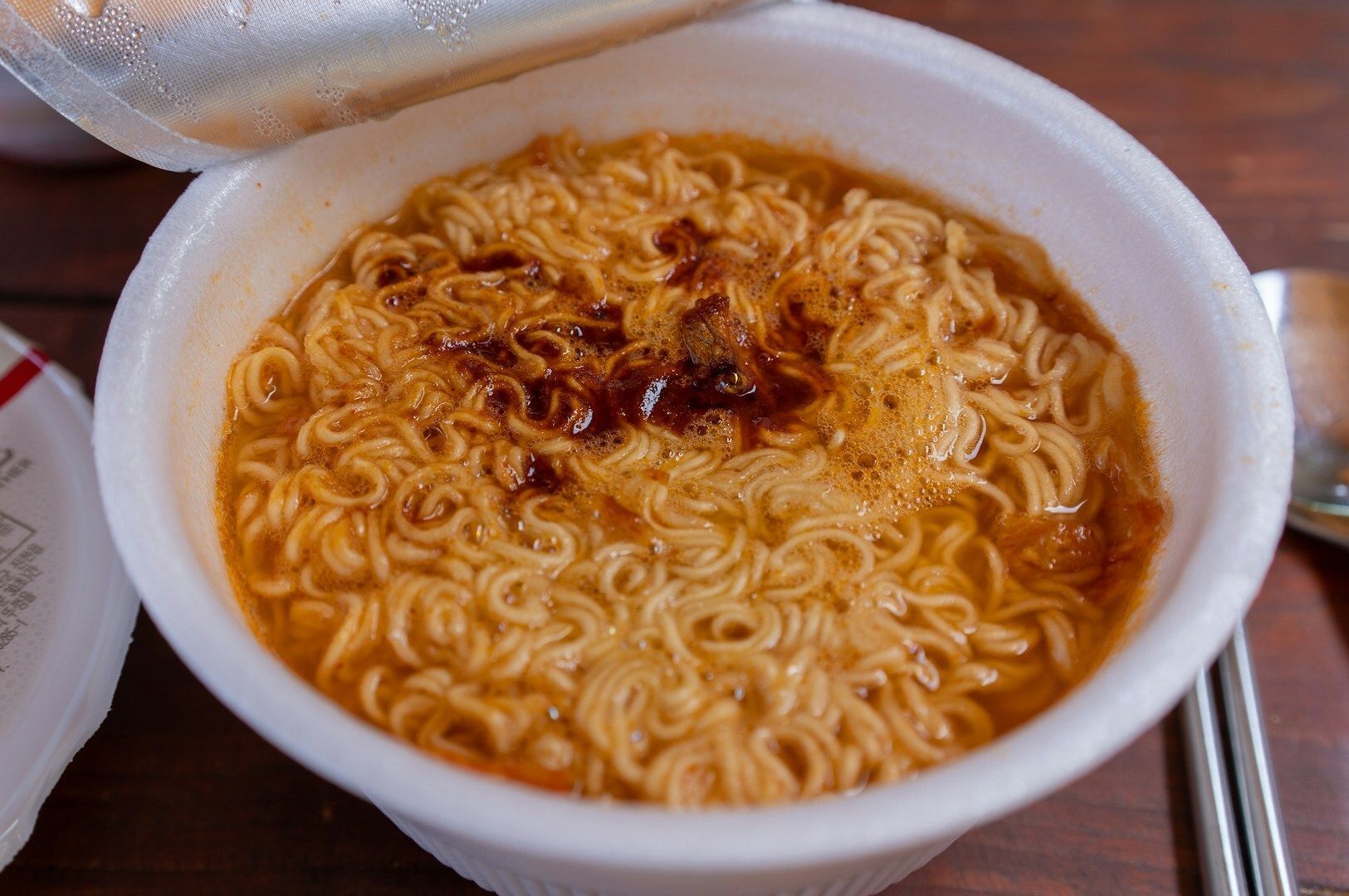 A bowl of Korean instant ramen with a spoon next to a can of soup, set on a rustic wooden surface.