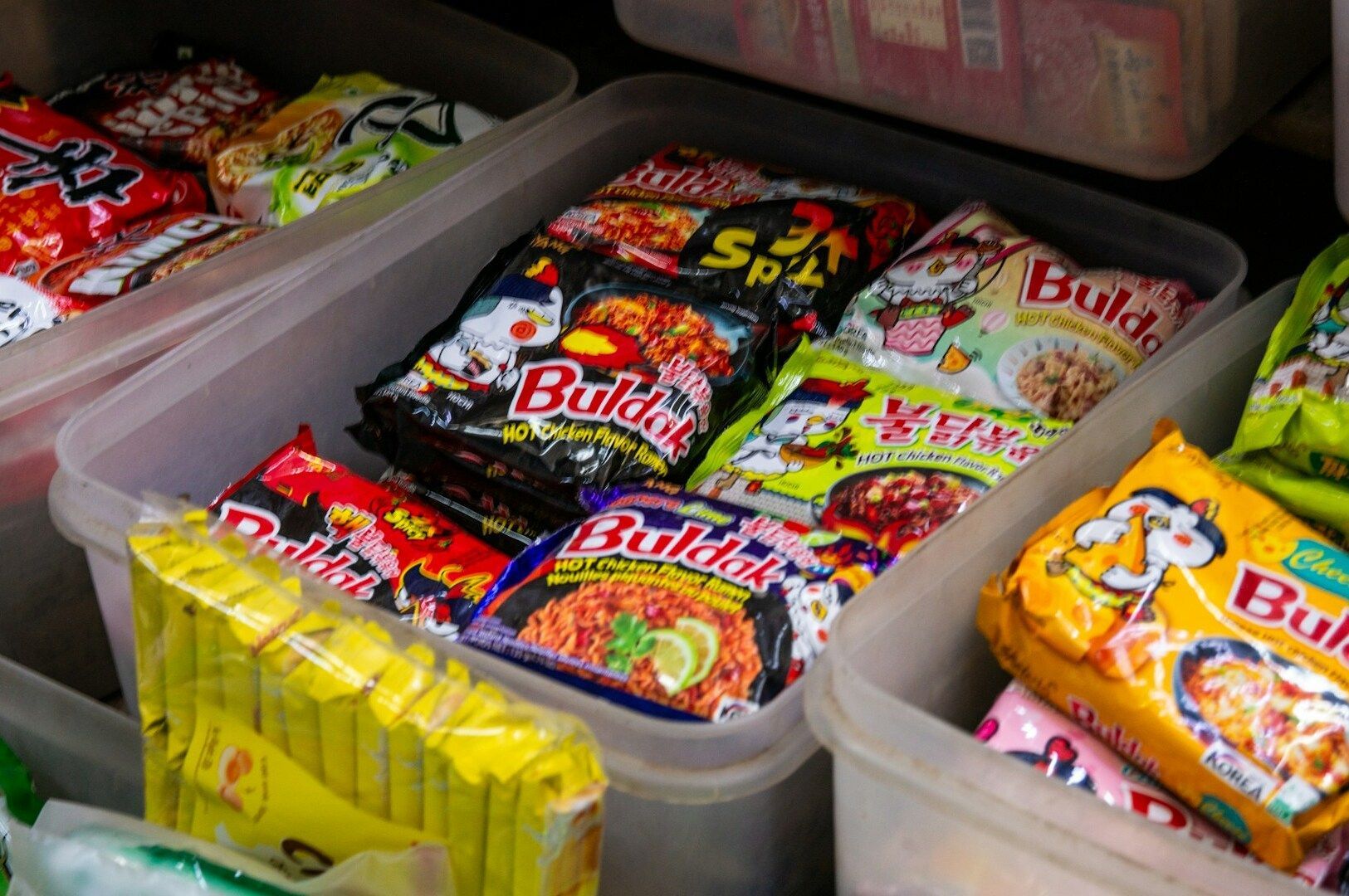 Assorted instant noodles, featuring Korean ramen or Buldak, arranged in transparent plastic containers for easy viewing.