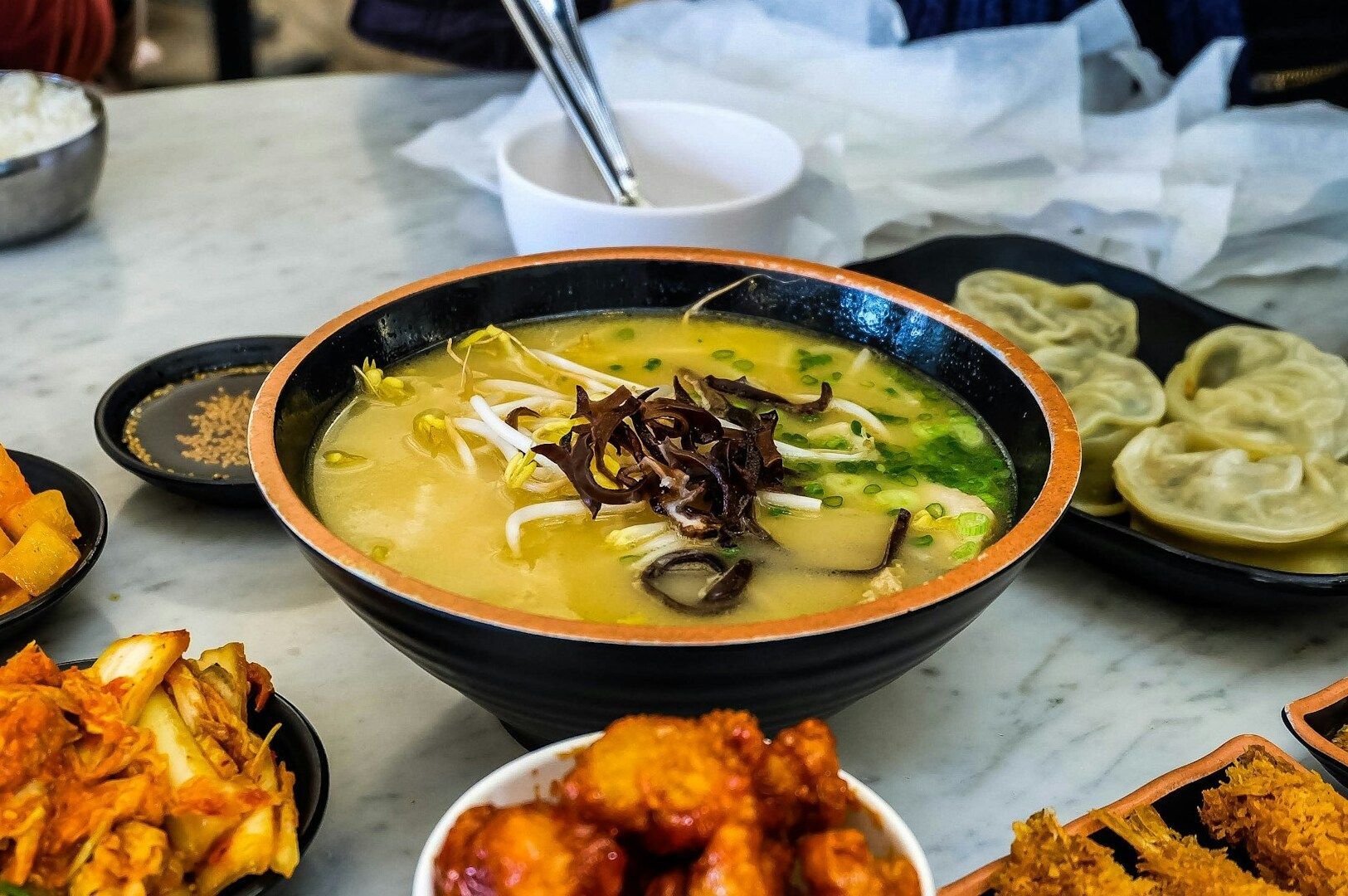 A bowl of Korean ramen featuring dumplings, placed on a table with an assortment of other foods.