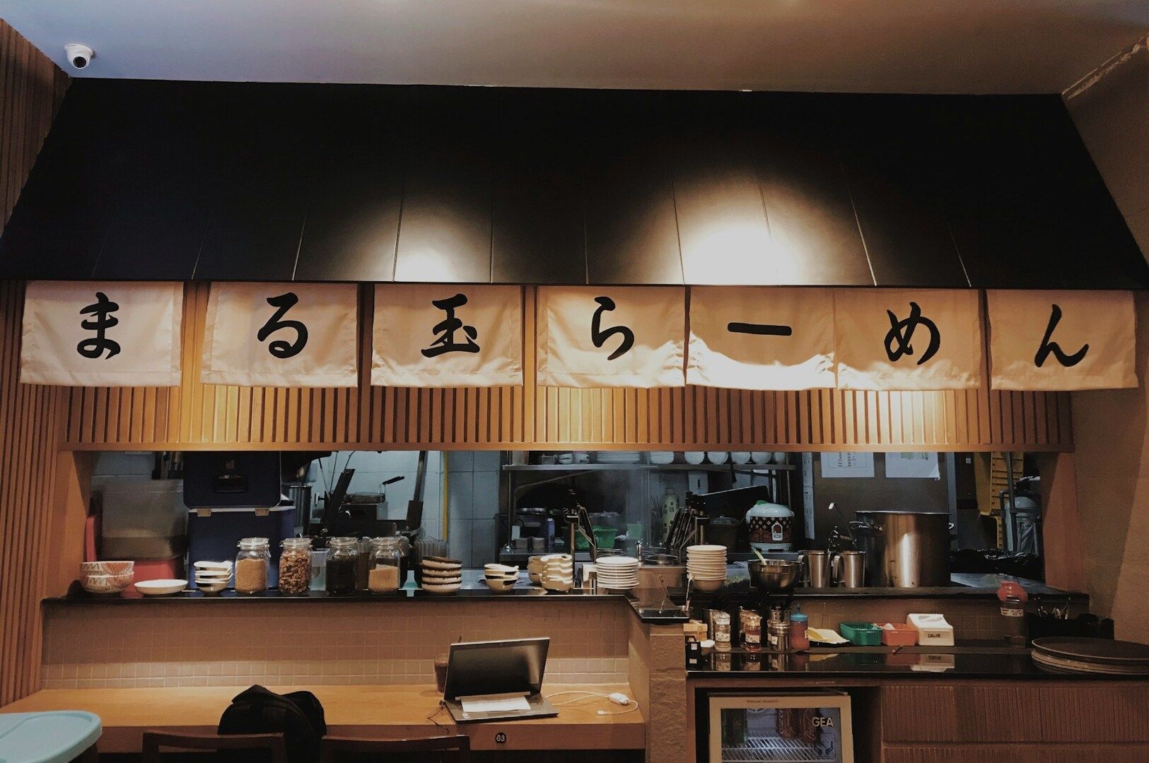 The image depicts the cozy interior of a small Japanese ramen shop, featuring a wooden counter. The warm atmosphere is enhanced by the aroma of ramen noodles and various toppings like soft boiled eggs and green onions, while steam gently rises into the air.