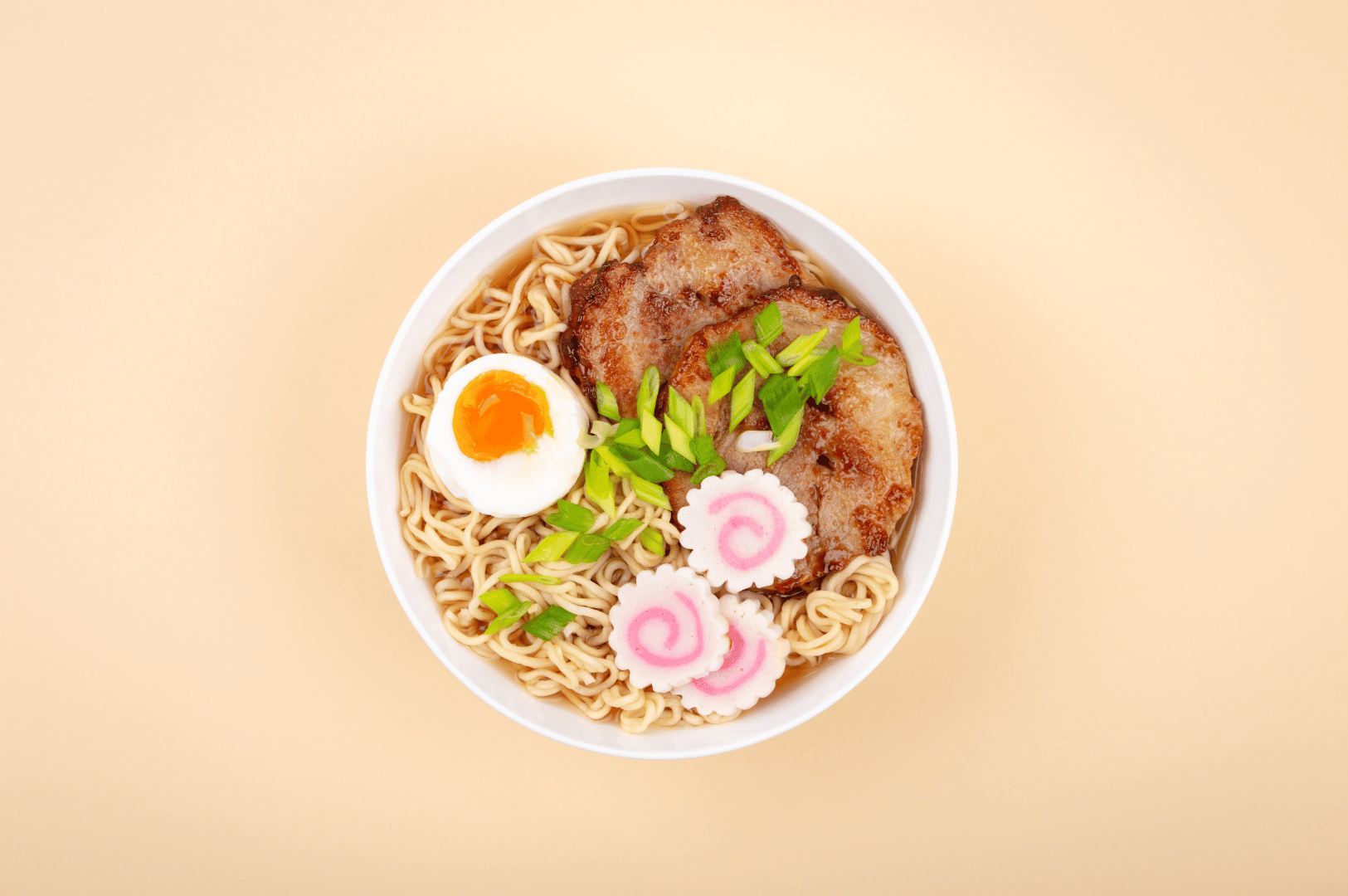 Steaming bowl of Japanese ramen topped with grilled pork slices, soft-boiled egg, green onions, and three narutomaki fish cakes with pink swirls, highlighting the iconic ramen garnish.