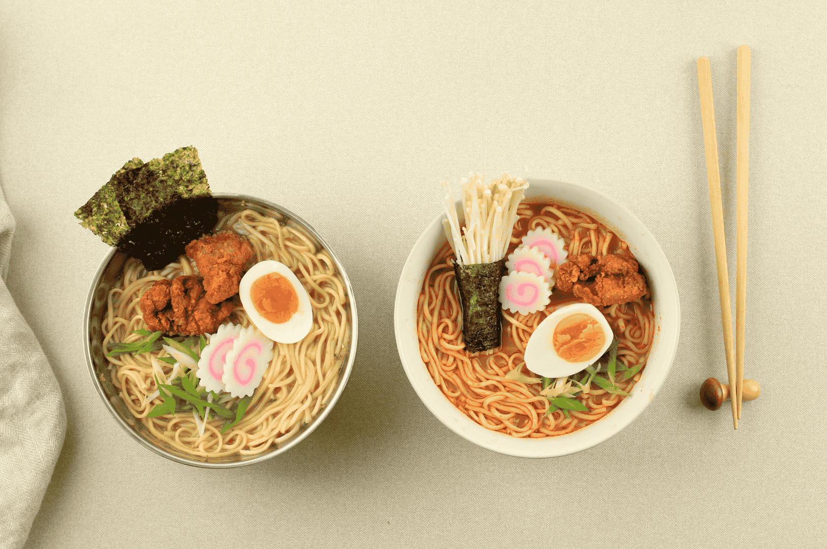 Two bowls of Japanese ramen on a light surface—one topped with fried chicken, narutomaki fish cakes, seaweed, green vegetables, and soft-boiled egg; the other with enoki mushrooms wrapped in seaweed alongside similar toppings—served with wooden chopsticks on a holder, showcasing the variety and artistry of ramen presentation.