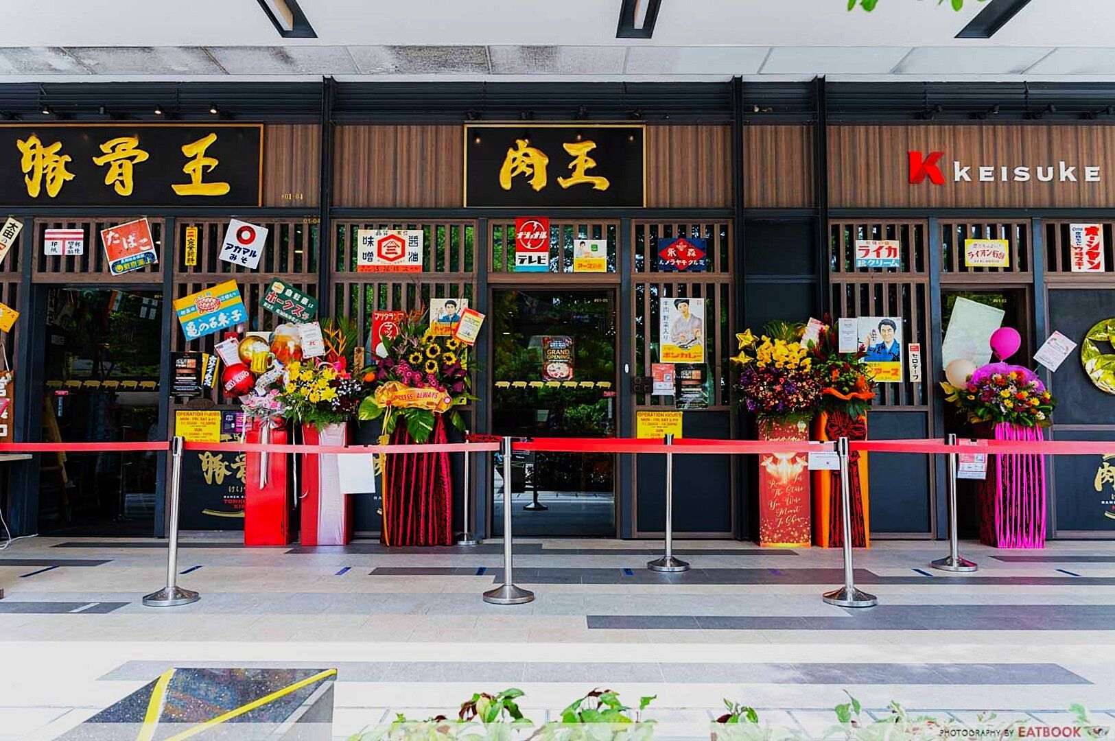 Storefront of a restaurant with Japanese signage, adorned with vibrant floral arrangements, balloons, posters, and ceremonial decorations. A red ribbon indicates an opening event.