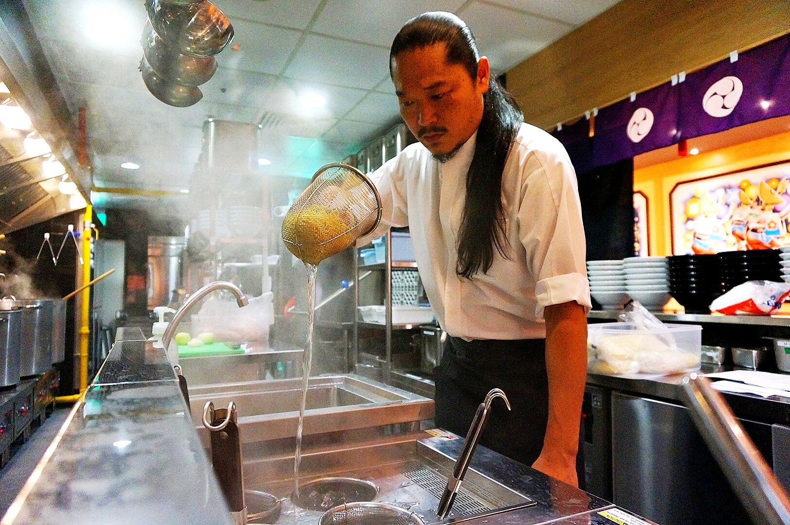 Focused chef Keisuke Takeda with long hair prepares noodles in a steamy kitchen, skillfully draining water with a mesh strainer. The environment is busy yet calm.