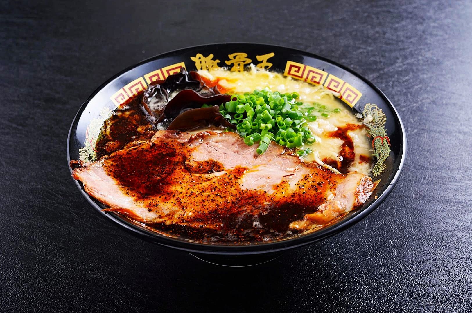 A black bowl filled with rich tonkotsu ramen, featuring a slice of roasted pork, dark soy sauce, green scallions, and a creamy broth.