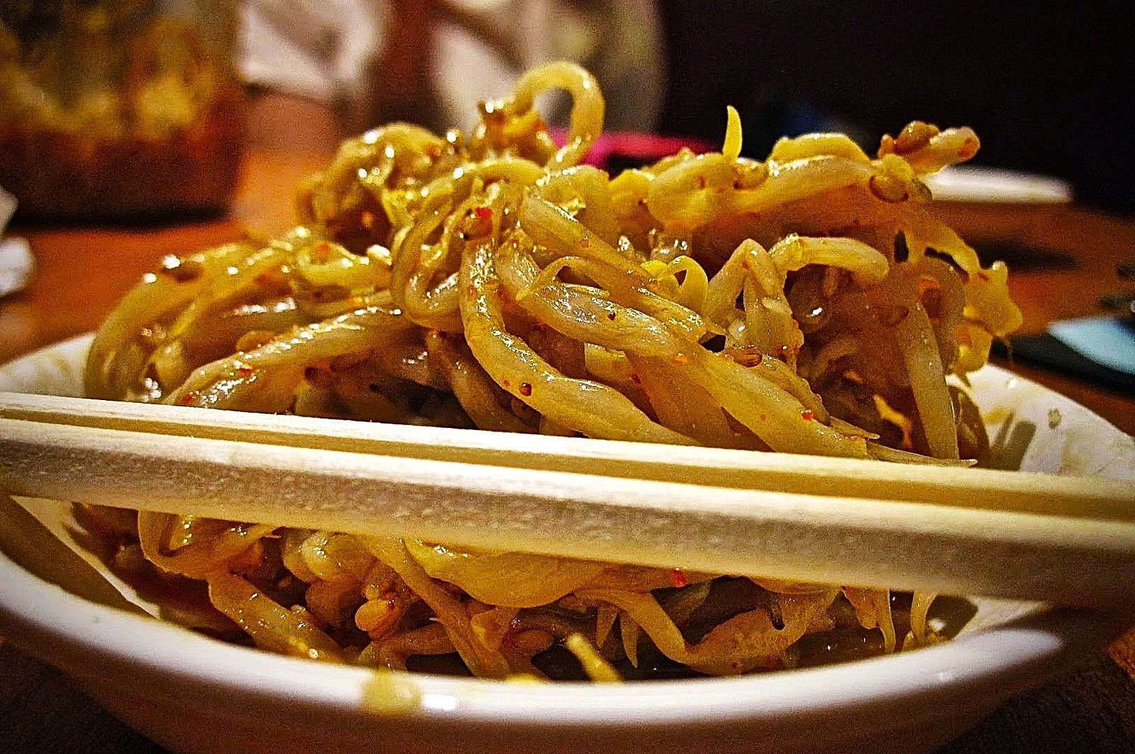 A close-up of stir-fried bean sprouts seasoned with spices on a white plate, with wooden chopsticks resting on top. The background is softly blurred.