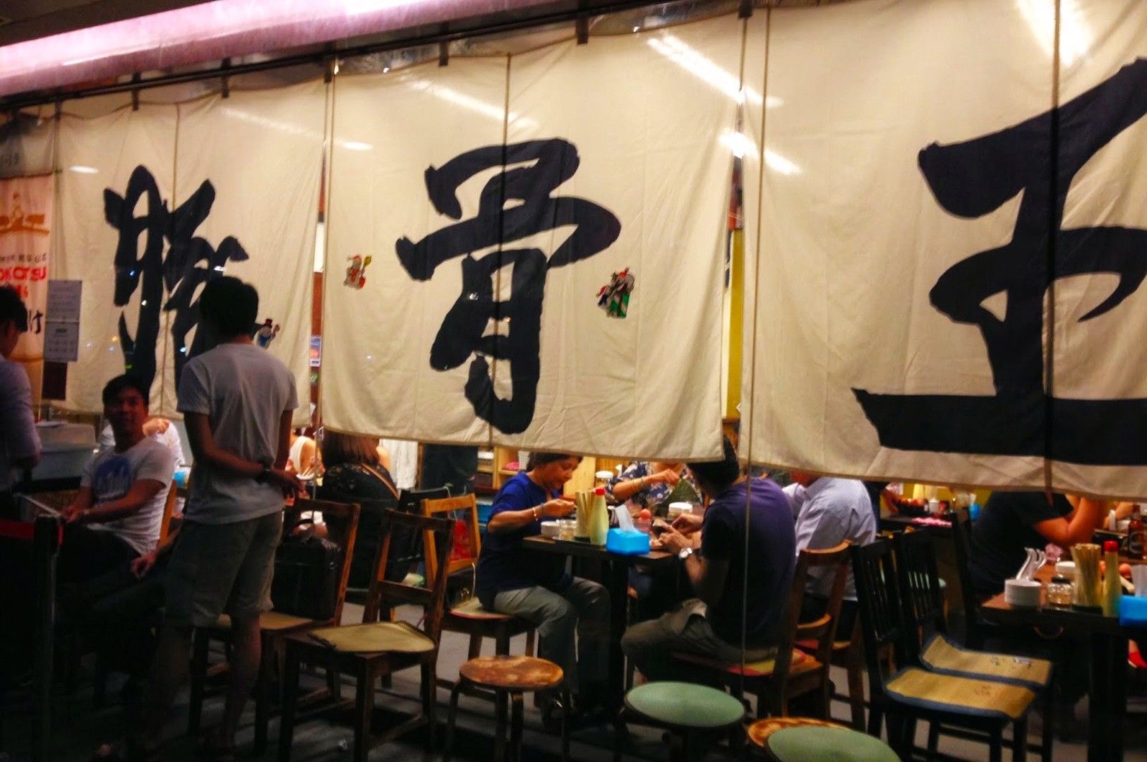 Dimly lit Ramen Keisuke Tonkotsu King restaurant at Tonkotsu King Orchid Hotel with people dining inside. Large cloth banners hang with bold Kanji characters. Cozy and bustling atmosphere.