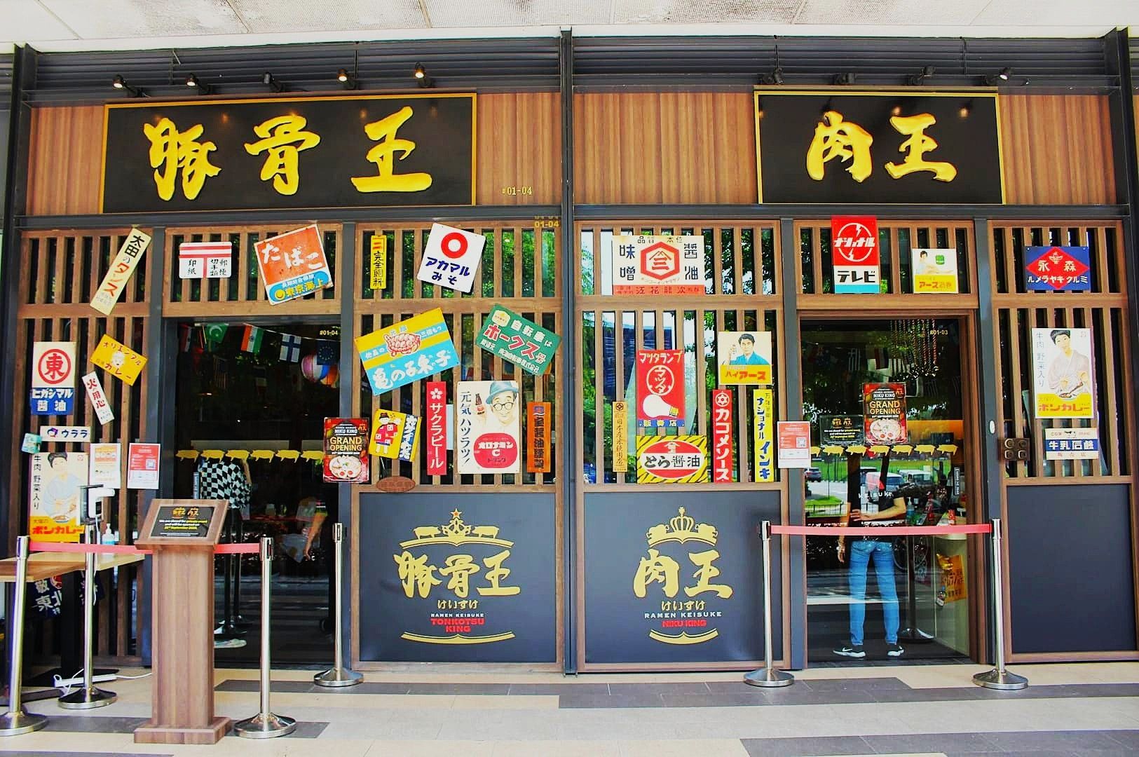 Front of a Keisuke Tonkotsu King restaurant with dark wood panels, bold gold signage, and colorful vintage posters on windows. Welcoming and lively atmosphere.
