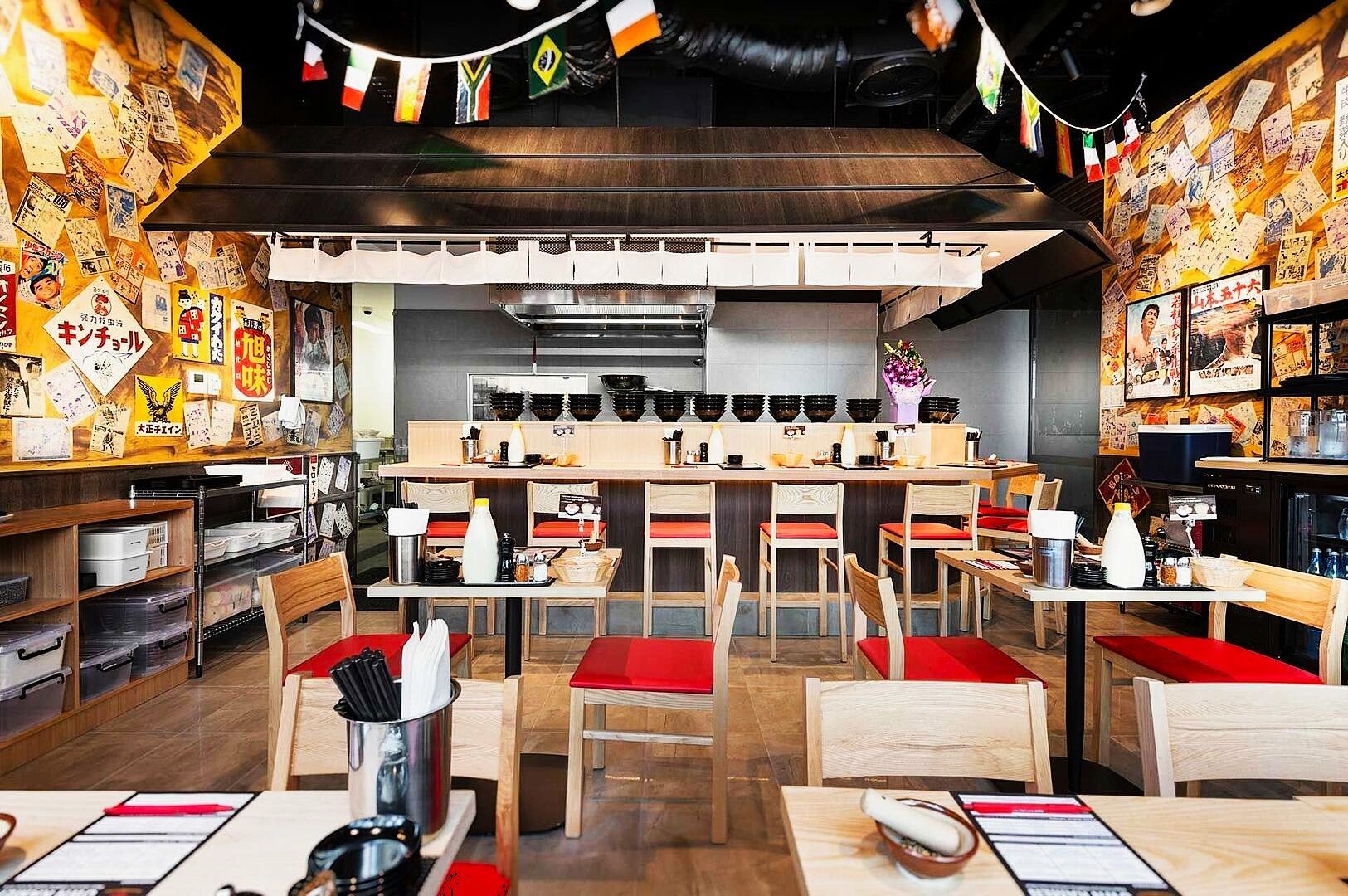 Ramen Keisuke Tonkotsu King restaurant features wooden tables with red cushions, a counter with black bowls, colorful wall posters, and hanging flags, creating a warm, inviting atmosphere.