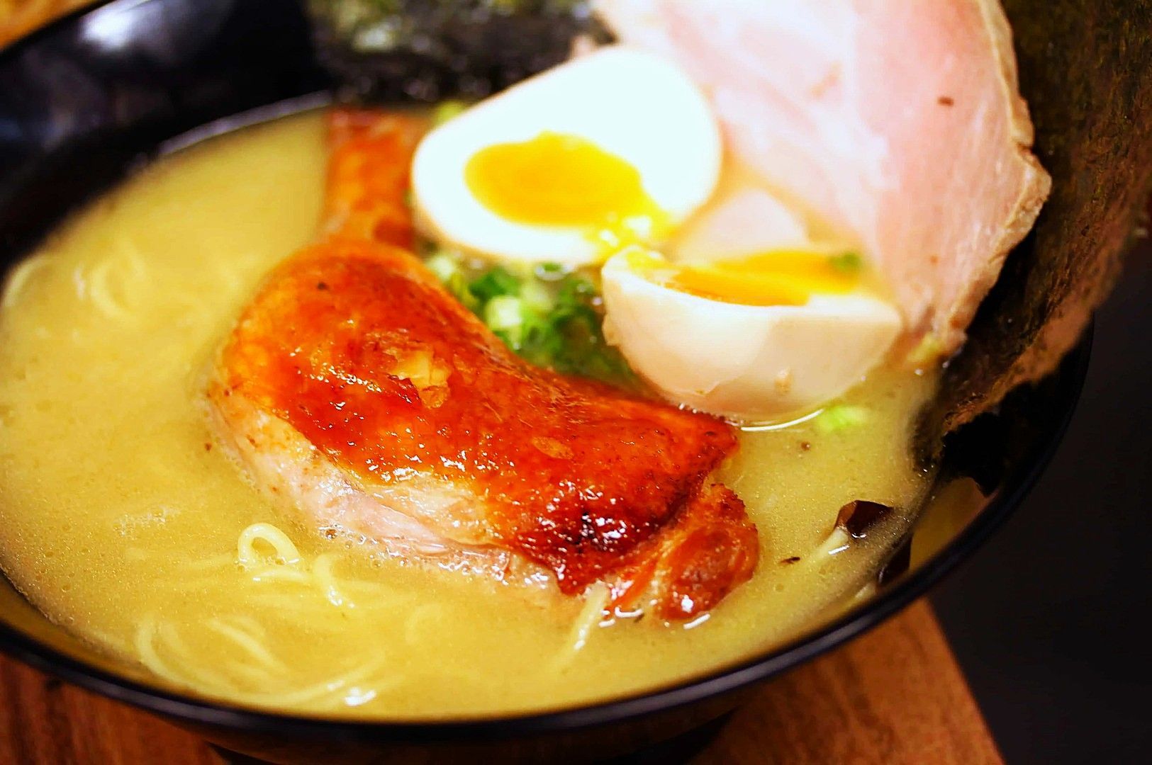 Close-up of a bowl of creamy ramen with sliced roasted chicken, half a soft-boiled egg, green onions, and seaweed. The dish appears savory and comforting.