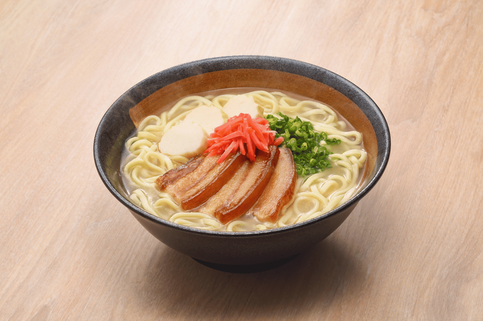 A steaming bowl of ramen in a dark ceramic vessel, layered with braised pork belly, fish cakes, scallions, and pickled red ginger over noodles in a light broth. Rising steam emphasizes freshness, while the rustic presentation reflects the artisanal, farm-to-table approach to Japanese cuisine celebrated in San Francisco.