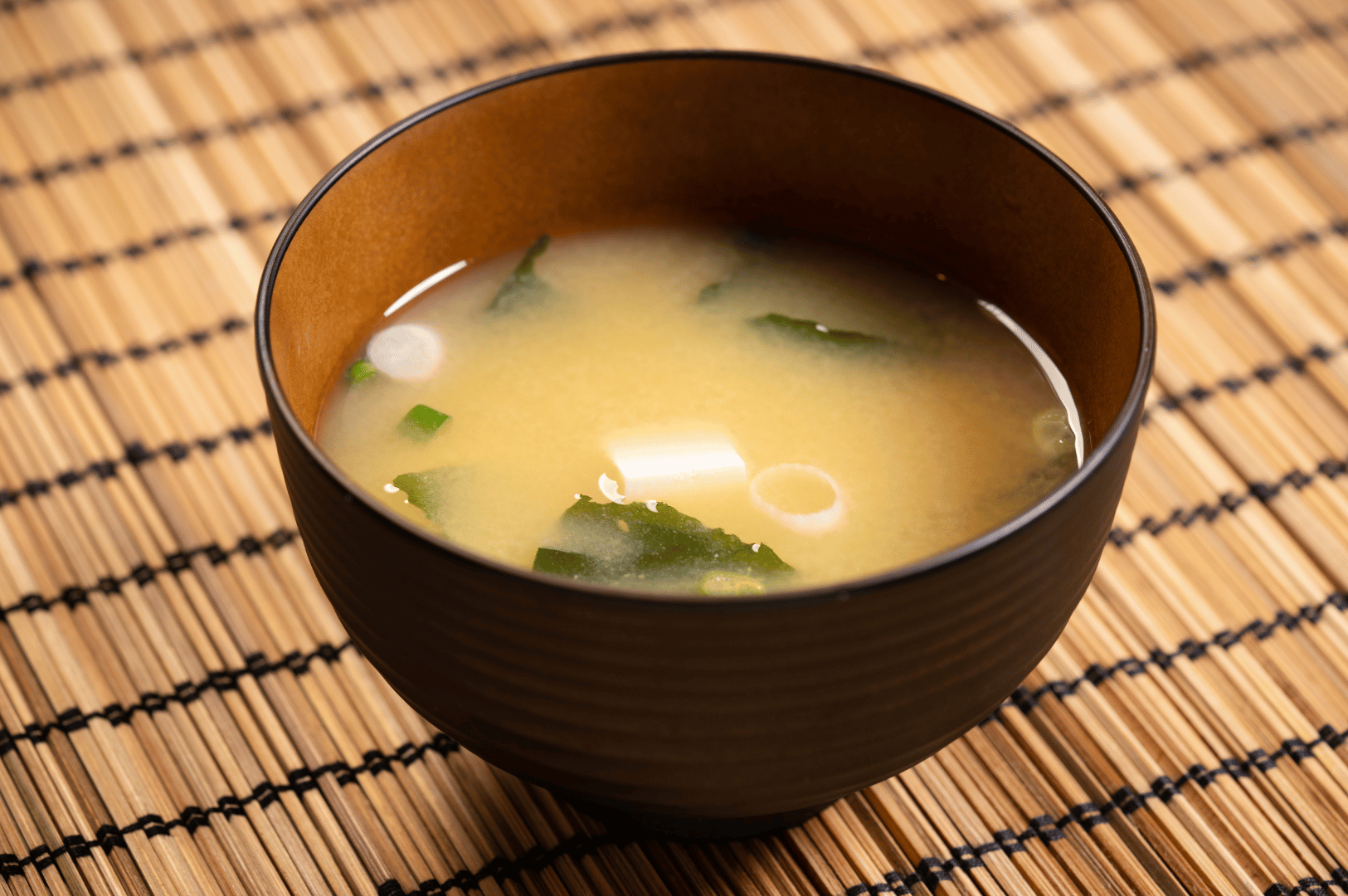 A traditional bowl of miso soup with tofu cubes, seaweed, and scallions, served on a bamboo mat. The dark ceramic vessel and natural textures emphasize simplicity and authenticity, reflecting the farm-to-table philosophy that connects Japanese culinary heritage with San Francisco’s artisanal dining culture.