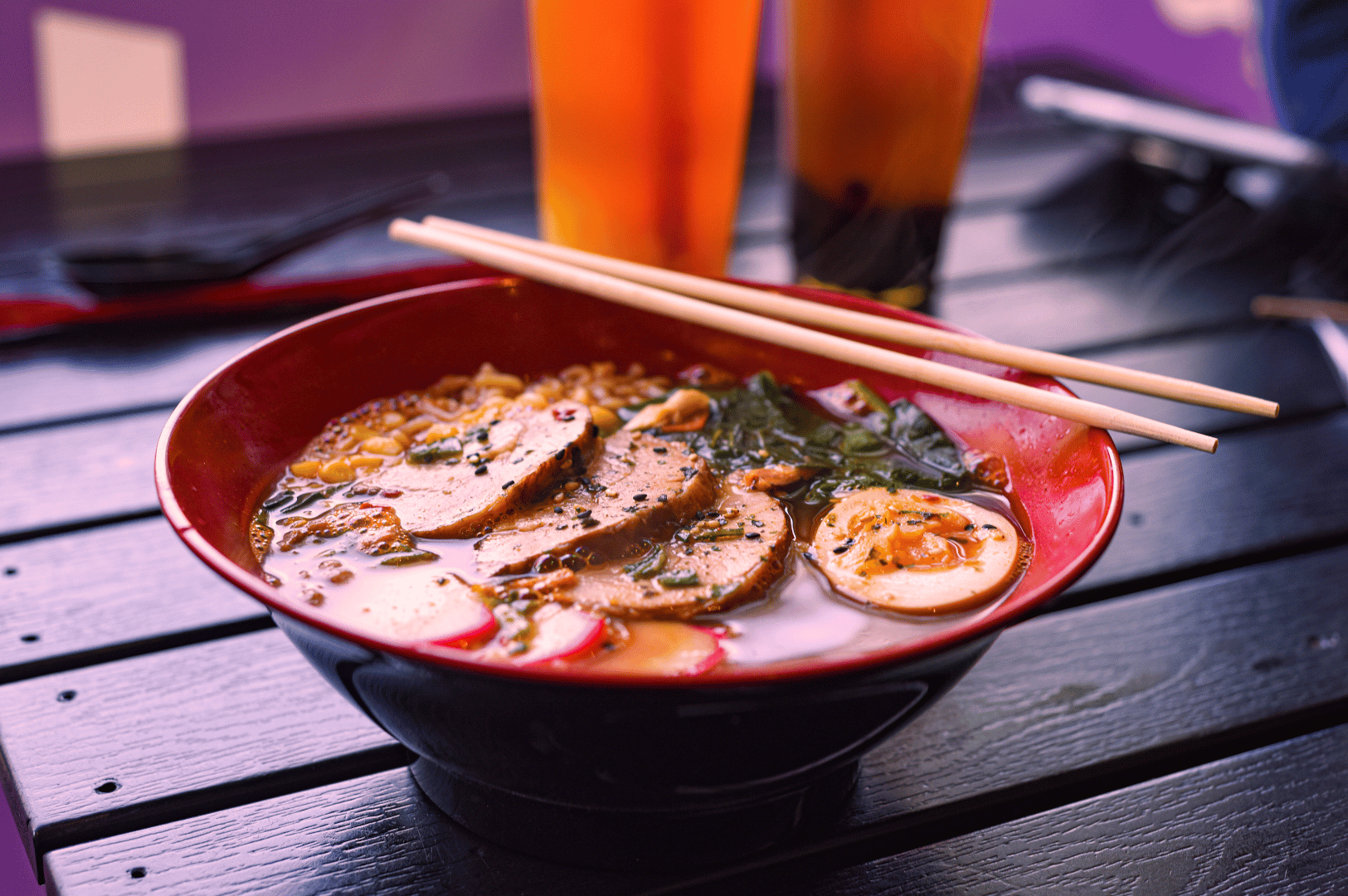 A vibrant bowl of ramen in a red and black vessel, layered with pork slices, a soft-boiled egg, leafy greens, and noodles in a rich broth. Chopsticks rest across the bowl, while iced drinks and a smartphone in the background evoke the modern dining culture where Japanese tradition meets San Francisco’s farm-to-table philosophy.
