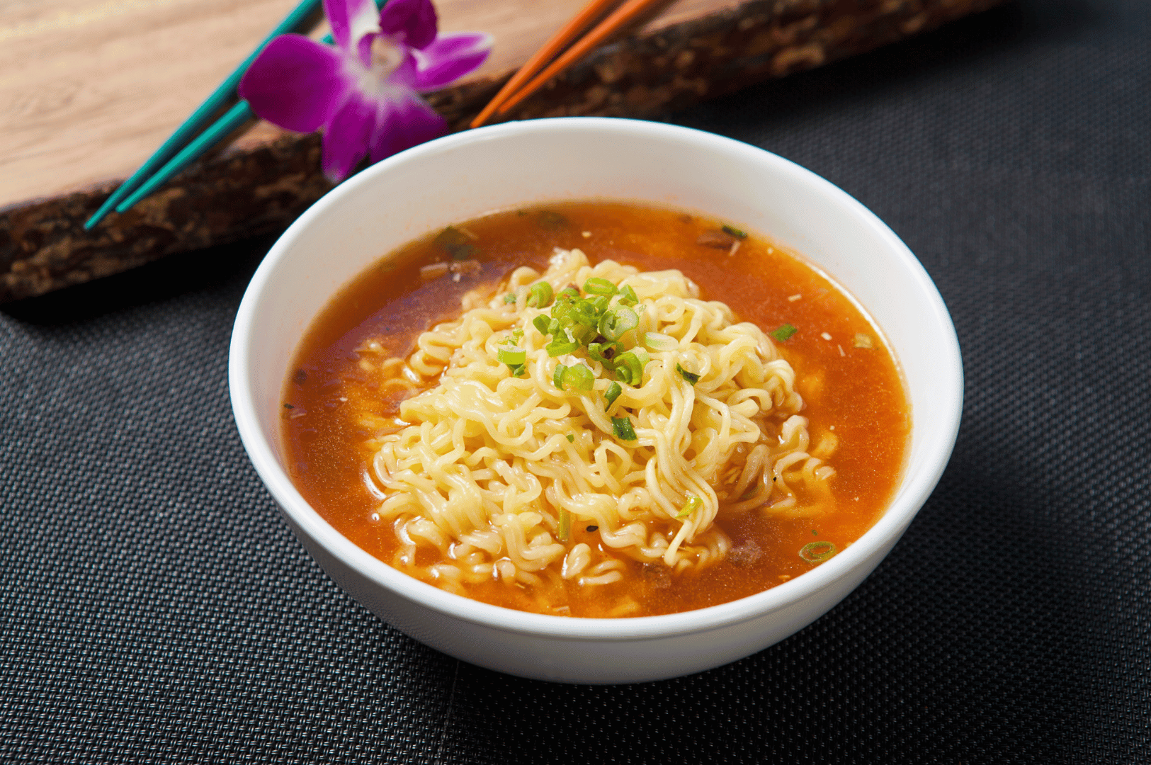 A bowl of instant ramen in a deep red broth, garnished with fresh green onions, sits on a dark textured surface. Brightly colored chopsticks and a purple orchid in the background add playful contrast and elegance—evoking the cozy, flavorful charm of Seoul’s noodle culture and the casual indulgence of a weekend ramen tour.