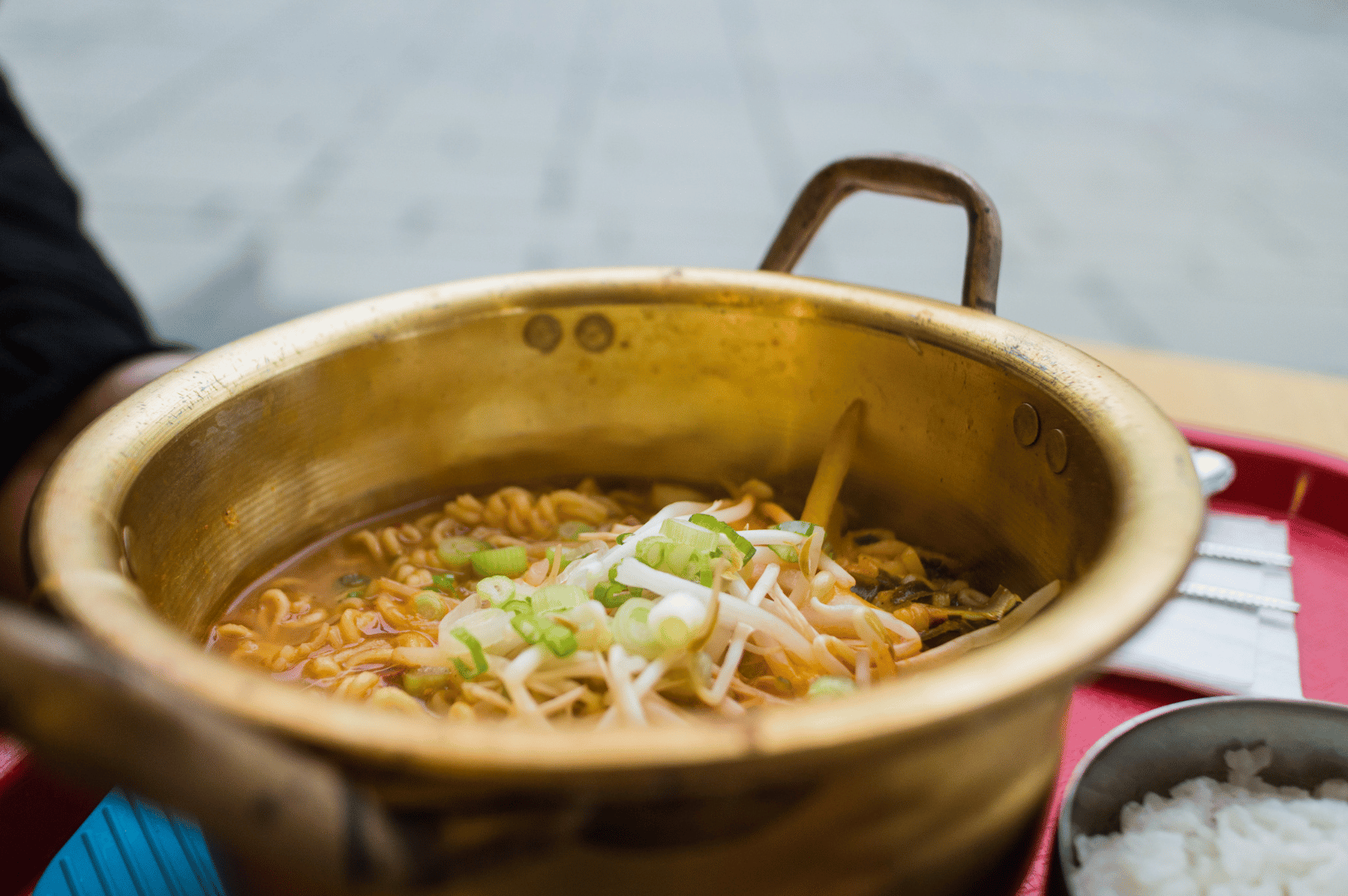 A brass pot of ramen served on a red tray, its rich broth topped with green onions and bean sprouts. A side of white rice in a metal bowl and neatly placed utensils evoke a comforting, everyday dining ritual—capturing the warmth and authenticity of Seoul’s noodle districts during a weekend ramen getaway.