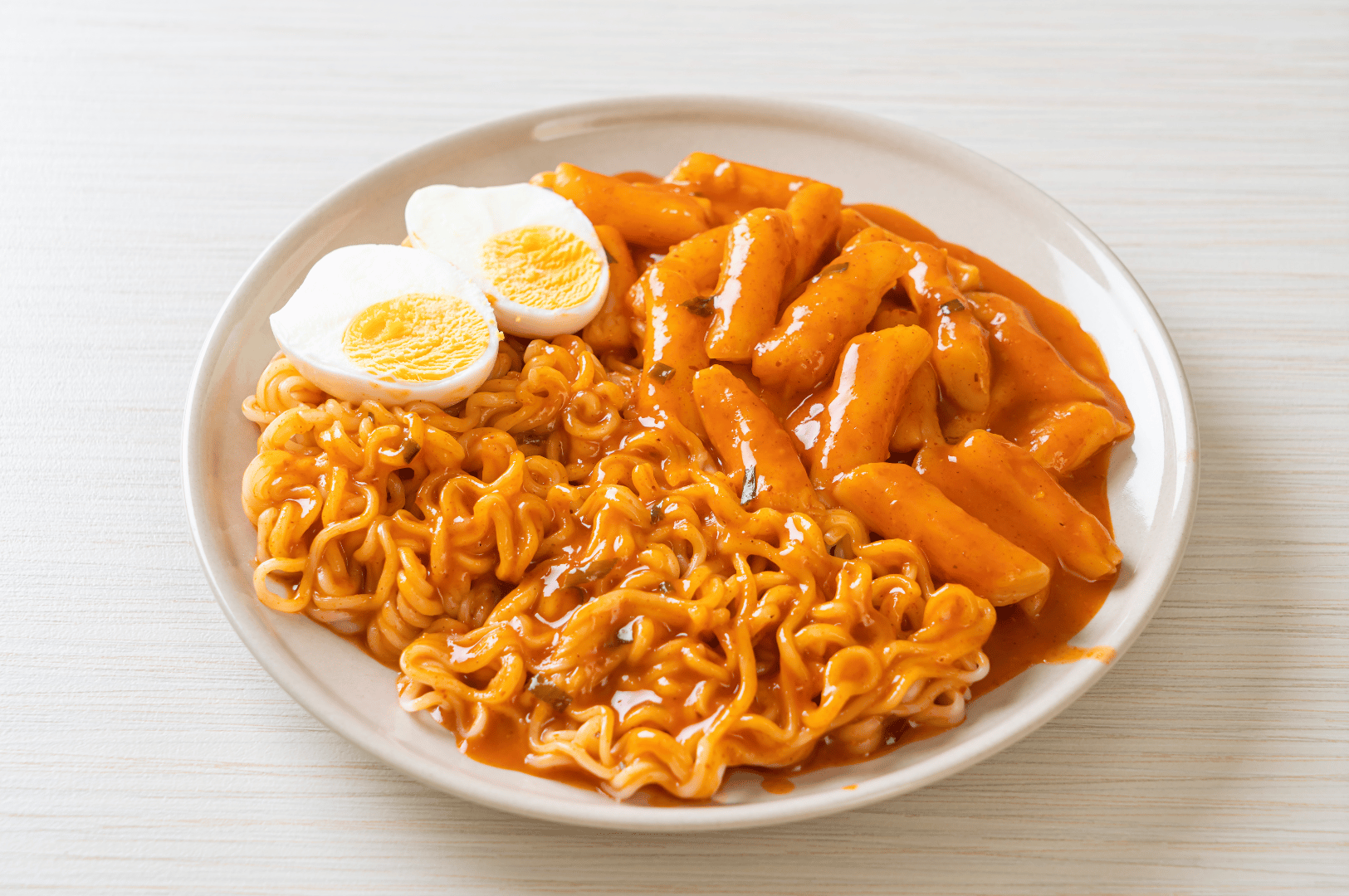 A plate of spicy tteokbokki paired with glossy ramen noodles and halved boiled egg, all drenched in a vibrant red-orange sauce. Set on a light wooden surface, the dish radiates warmth and indulgence—blending street food comfort with bold flavor, a perfect stop on Seoul’s weekend ramen tour through its noodle-loving districts.
