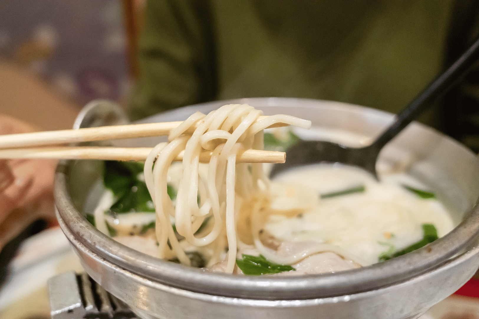 A close-up of creamy tonkotsu ramen served in a metallic bowl, with noodles lifted mid-air by wooden chopsticks, revealing their silky texture. Green vegetables and tender meat pieces float in the milky broth, while a black spoon rests inside—evoking the warmth and tactile pleasure of a dish born from hours of bone simmering and emulsification.