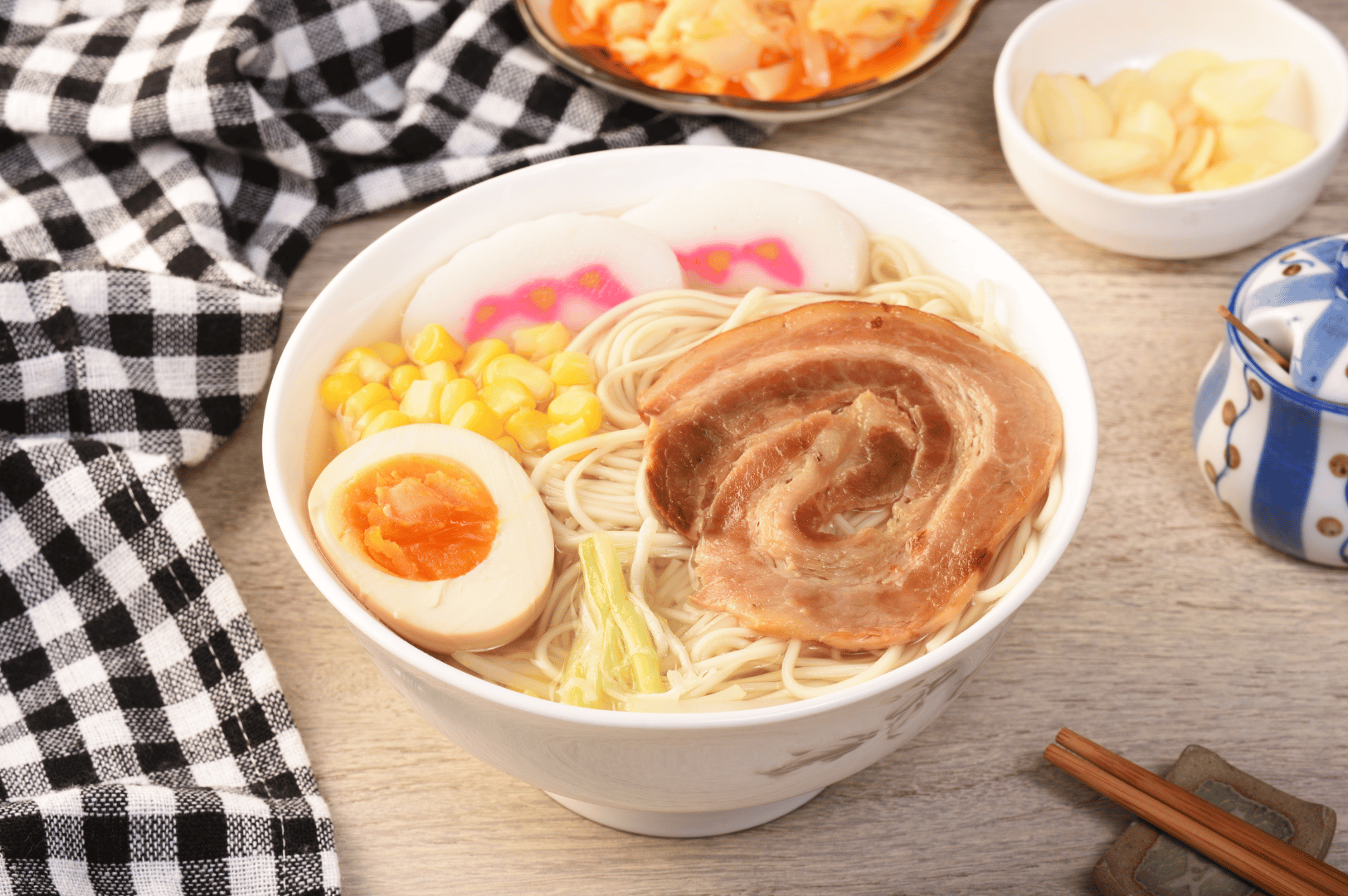 A richly garnished bowl of shio ramen sits on a wooden table, topped with braised chashu, molten-yolk egg, sweet corn, narutomaki fish cakes, and shredded green onions. Surrounding side dishes—pickled vegetables, potato chips, and a ceramic condiment jar—add texture and cultural depth, while a checkered cloth and resting chopsticks evoke the warmth of a home-cooked feast born from scientific precision and soulful tradition.